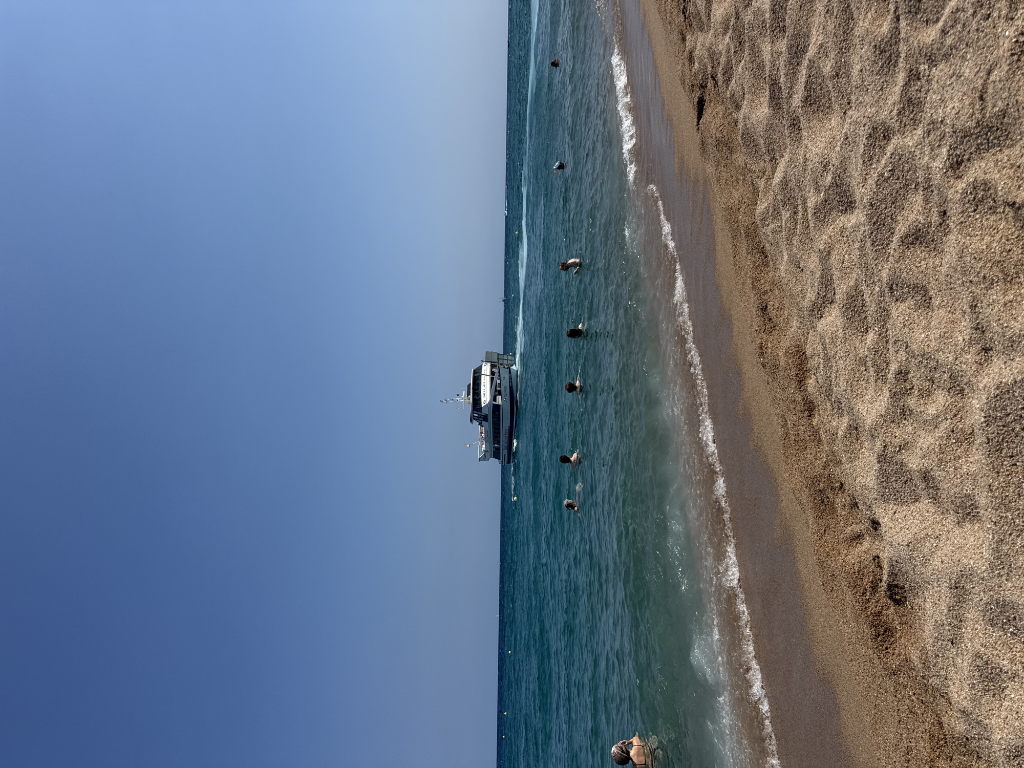 This is a landscape photograph of a beach in Blanes, Spain, on a clear, sunny day. The foreground is dominated by a sandy beach with rippled textures caused by wind or footprints. The middle ground features the turquoise Mediterranean Sea, with gentle waves lapping at the shore. Several people are visible in the water, swimming and enjoying the sea. Further out in the sea, a white passenger boat with multiple decks is sailing. In the background, the horizon is a sharp line between the sea and a cloudless, bright blue sky. The scene captures a typical summer day at a coastal resort.
