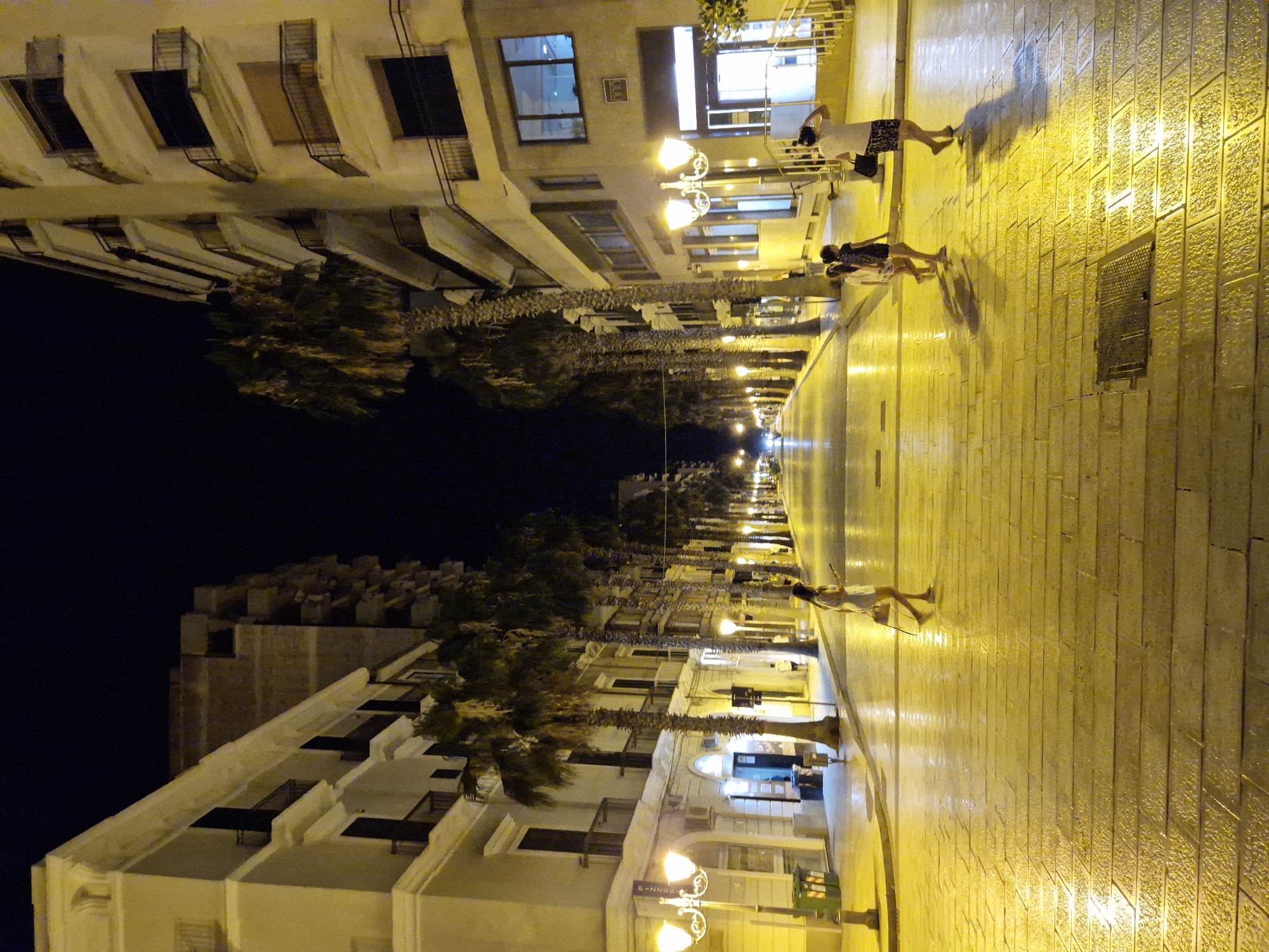The image depicts a street scene at night in the city of Taranto, Italy, likely on Via Regina Margherita or a similar pedestrianized street lined with palm trees. The street is illuminated by streetlights, casting a warm, yellow glow on the wet pavement, which is paved with large rectangular stones. Buildings with multiple stories and balconies line both sides of the street. Several palm trees are visible, their fronds silhouetted against the dark sky.

In the foreground, a woman in a white dress walks from left to right, holding a bag over her shoulder and her phone. Further down the street, a man in a white t-shirt and patterned shorts walks away from the camera towards the right, with a black bag in his left hand. Two young girls, one in a dark patterned dress and the other in a white top and shorts, walk beside him, also moving away from the camera.

The street appears relatively empty, with the figures of people walking and the ambient light creating a sense of calm. The overall impression is one of a pleasant evening stroll in a well-lit urban environment.