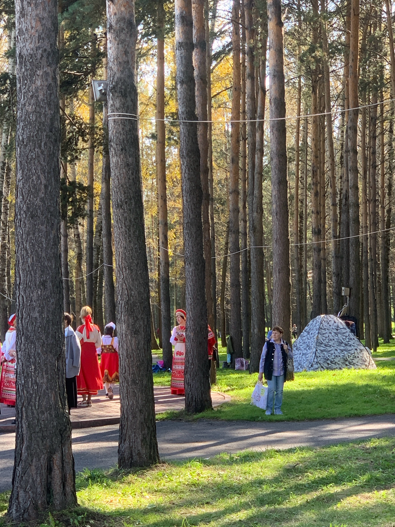 The image depicts a forested area, likely a park, with tall pine trees. Several people are present. On the left side of the frame, a group of individuals, some wearing traditional red and white embroidered clothing, are gathered. Further to the right, a person in casual clothing, a blue vest over a plaid shirt and jeans, stands holding two shopping bags. A camouflage-patterned tent is visible in the background to the right. The ground is covered in grass and a paved path runs through the scene. Sunlight filters through the trees, casting shadows on the grass. The overall atmosphere suggests a daytime outdoor gathering or event. The location is specified as Prokopyevsk, Russia.