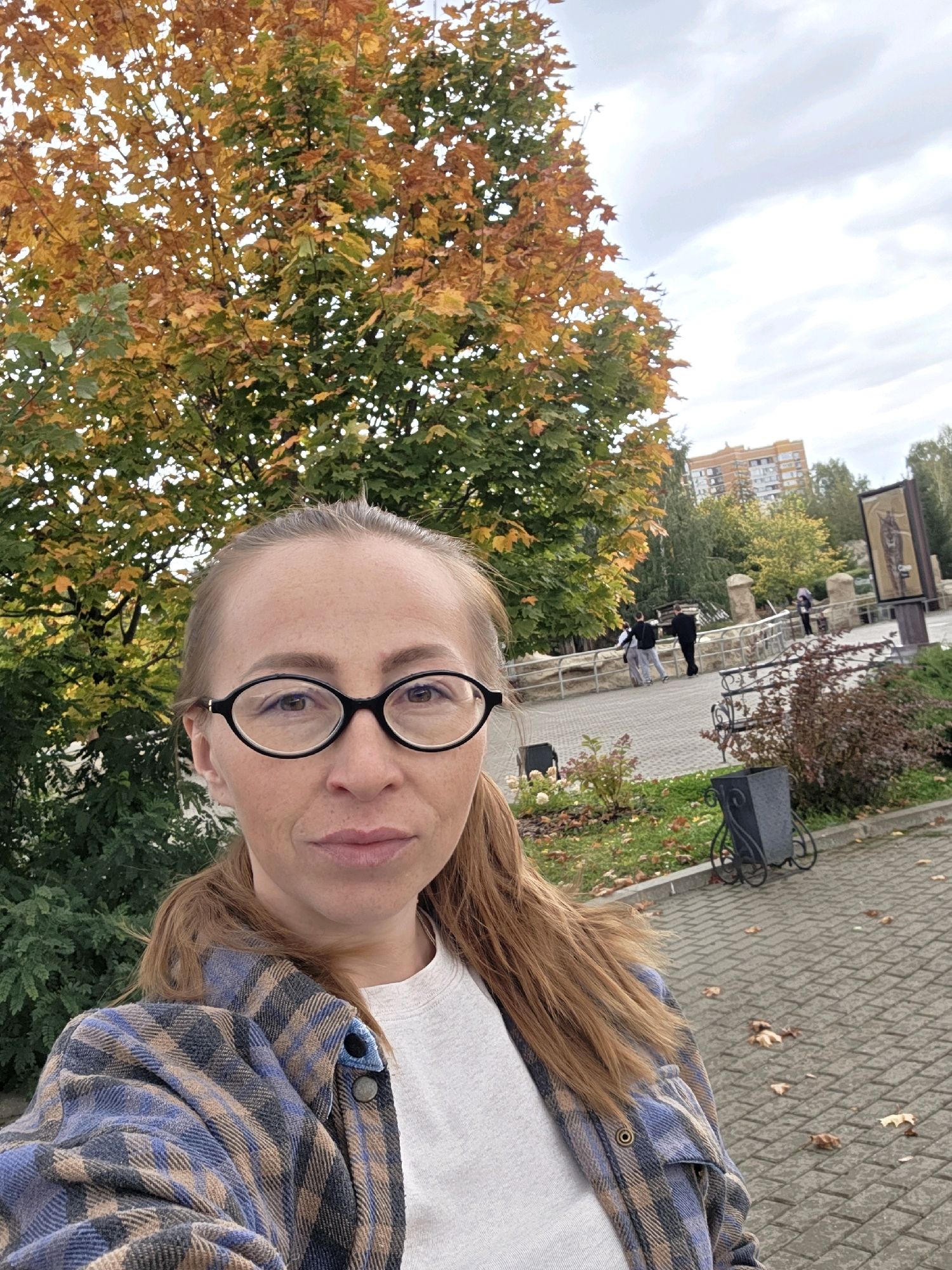 The image is a selfie taken outdoors in Izhevsk, Russia, during autumn. The foreground features a woman with fair skin and auburn hair, wearing glasses and a plaid shirt over a white t-shirt. She is looking directly at the camera. Behind her, a large tree displays colorful autumn foliage, with leaves in shades of yellow, orange, and green. In the background, a paved walkway is visible, with fallen leaves scattered on the ground. Several people are walking in the distance, and a multi-story building with an orange facade is visible in the far background. The sky is overcast.