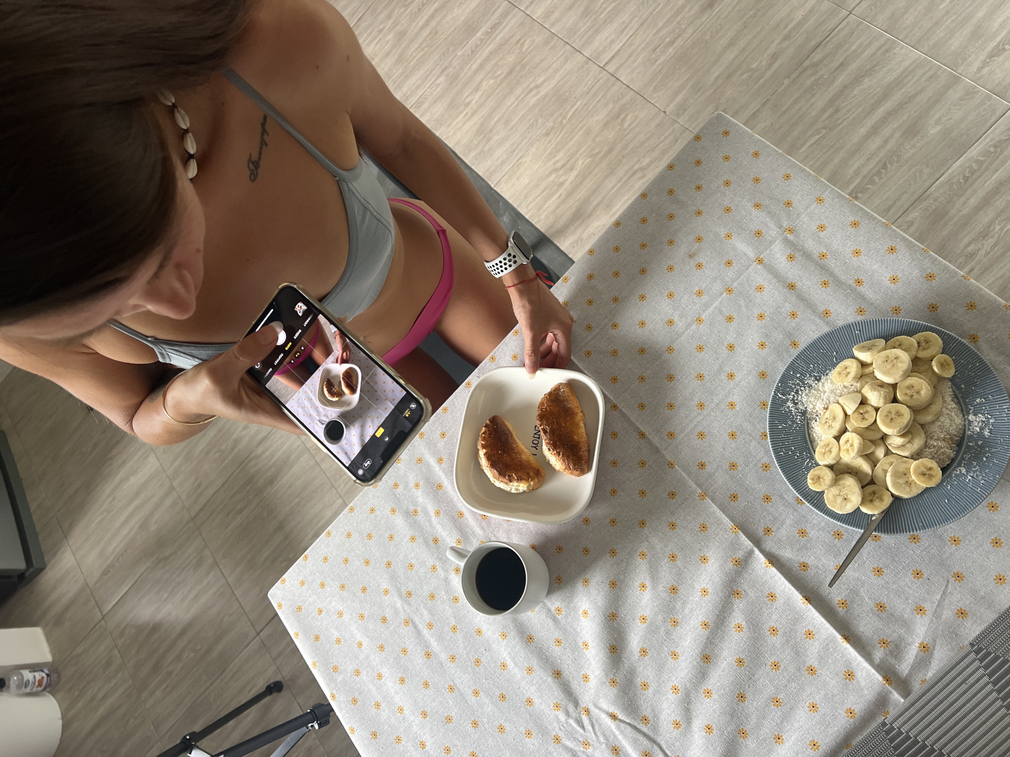 A woman is taking a photograph of breakfast with her smartphone. The breakfast consists of two croissants and a cup of coffee on a white plate with a patterned tablecloth. In the foreground, there is another plate with sliced bananas and shredded coconut, with a spoon resting on it. The woman is wearing a blue bikini top and pink bottoms. She has a seashell necklace and a watch on her left wrist. The scene appears to be in a kitchen or dining area with tiled flooring. The location is not visible in the image.