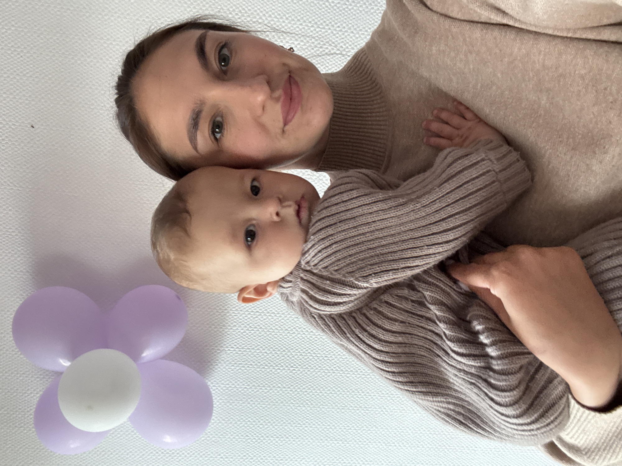A woman holds a baby. Both are wearing beige sweaters. A flower made of purple and white balloons is visible in the lower left corner. The location cannot be determined from the image.