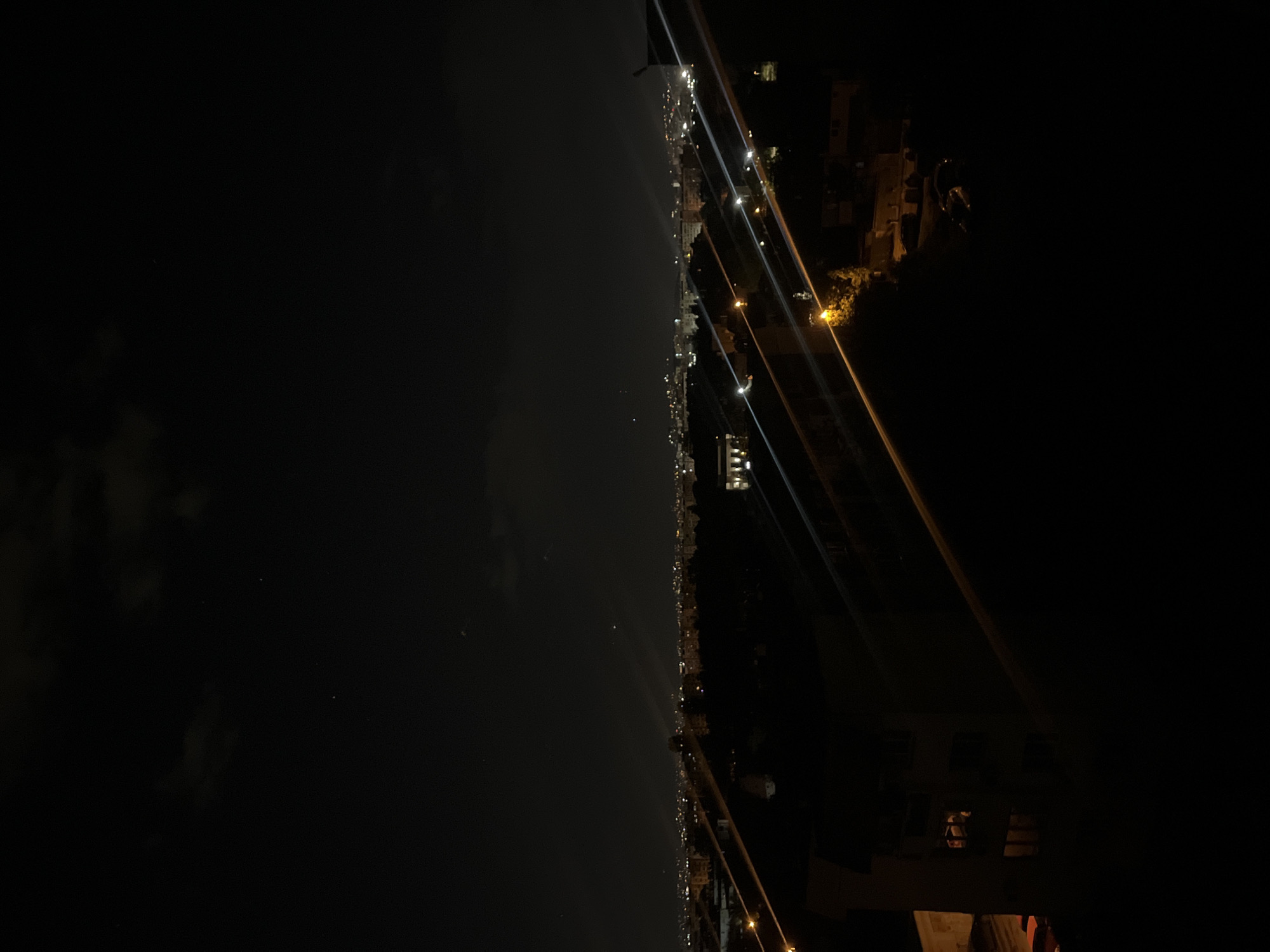 The image is taken at night in what appears to be Sofia, Bulgaria, showing a view of the city skyline with numerous lights extending along the horizon. The left side of the image is predominantly dark, suggesting either a body of water or a dimly lit area. The right side contains a few buildings and infrastructure elements, with visible wires or ropes stretching across the view. Illumination from streetlights and buildings contributes to the city's glow. There are no clearly discernible people or specific actions visible in the image. The overall environment is dark, with the city lights providing the primary source of illumination.
