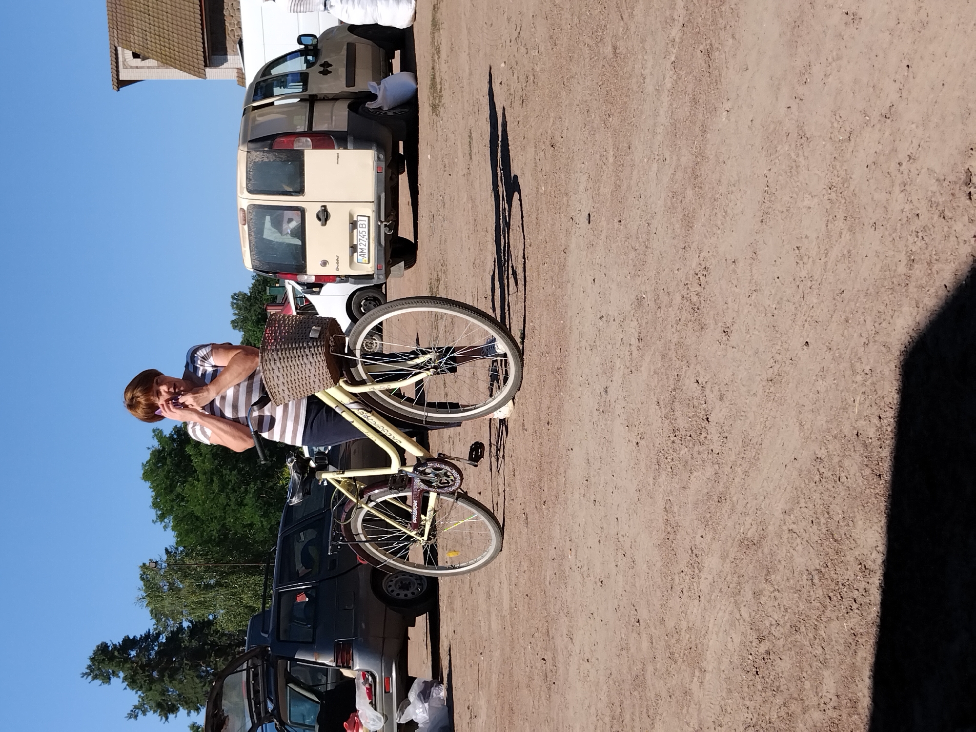 A woman with short brown hair, wearing a striped t-shirt and dark pants, is standing next to a light yellow bicycle on a dirt ground. She is holding a purple mobile phone to her right ear and appears to be talking on it. The bicycle has a wicker basket on the handlebars and a rack over the rear wheel. In the background, a dark grey car with its trunk open is parked on the left, and a beige van with its rear doors open is parked on the right. The environment is an outdoor area with a clear blue sky overhead. The scene appears to be in Irshansk, Ukraine, given the vehicle license plate which appears to be Ukrainian.
