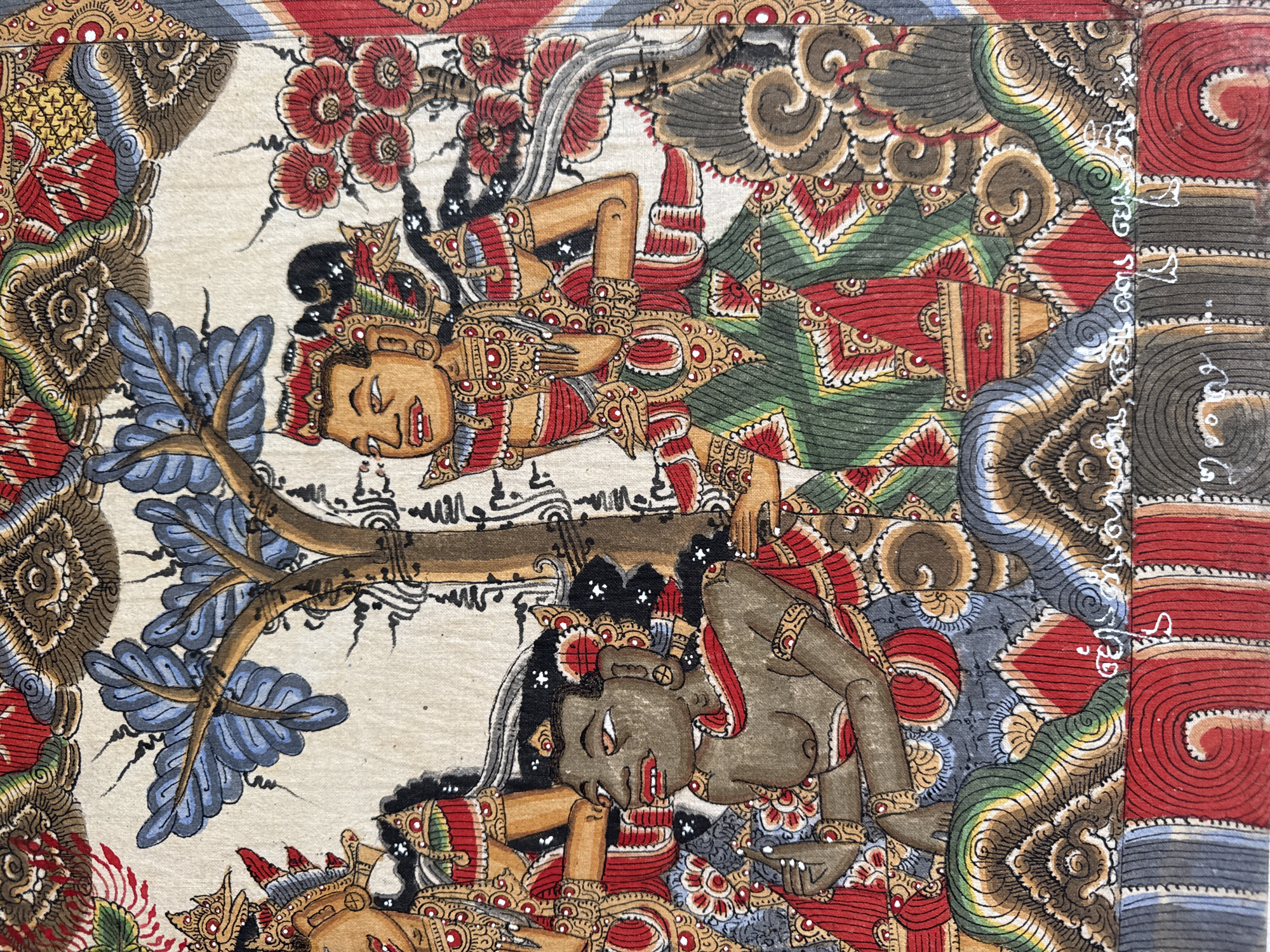 This image is a close-up of a traditional Balinese painting on fabric. The painting depicts two figures in a stylized, decorative manner, possibly from a mythological or religious narrative. The figures are depicted with brown skin tones, elaborate headdresses, and intricately patterned clothing in red, green, gold, and blue. One figure is lying down, with their head resting on their arm, appearing serene or possibly asleep. The other figure is also reclined, with their head tilted upwards and their mouth open, perhaps in a shout or song. They are positioned amidst swirling, organic patterns that suggest foliage, flowers, and clouds, rendered in a vibrant and detailed style. The background is a light, neutral color, highlighting the richness of the figures and decorative elements. The overall impression is one of movement and dynamism within a richly ornamented artistic tradition, likely originating from Bali, Indonesia, given the mention of Kuta Selatan. The painting appears to be a detail from a larger work. There is some visible white text, in a script that appears to be Balinese or Javanese, along the right edge of the image, integrated into the decorative border.