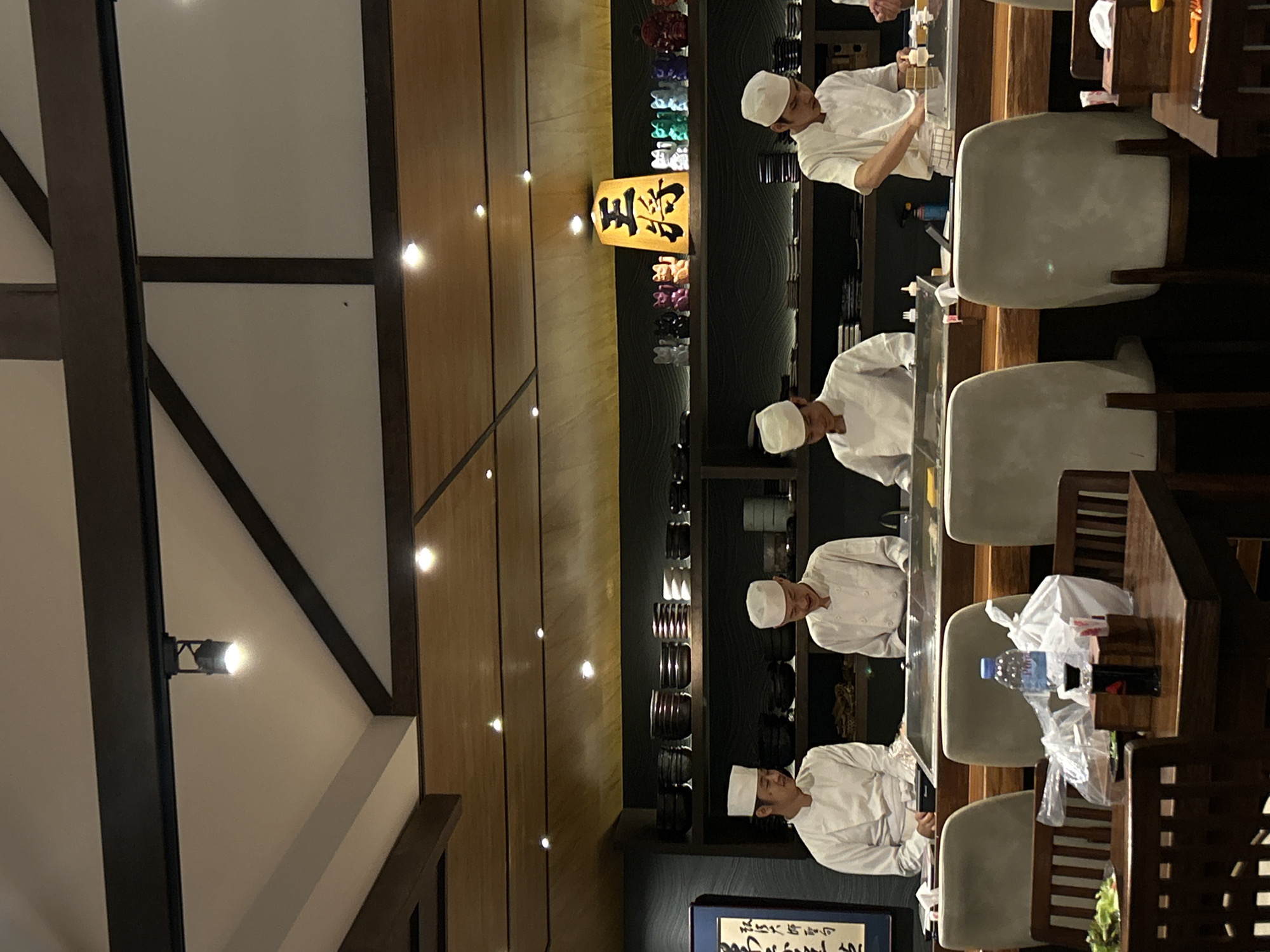 The image shows the interior of a restaurant in San Sai Luang, Thailand. Four chefs wearing white uniforms and hats are standing behind a counter preparing food. The chefs are positioned in a row. The restaurant features a light-colored ceiling with dark wooden beams. There are decorative items on display behind the chefs. Tables and chairs for patrons are in the foreground, with plastic bags and a water bottle visible on one of the tables. A decorative sign is visible on the wall. The lighting in the restaurant is warm.