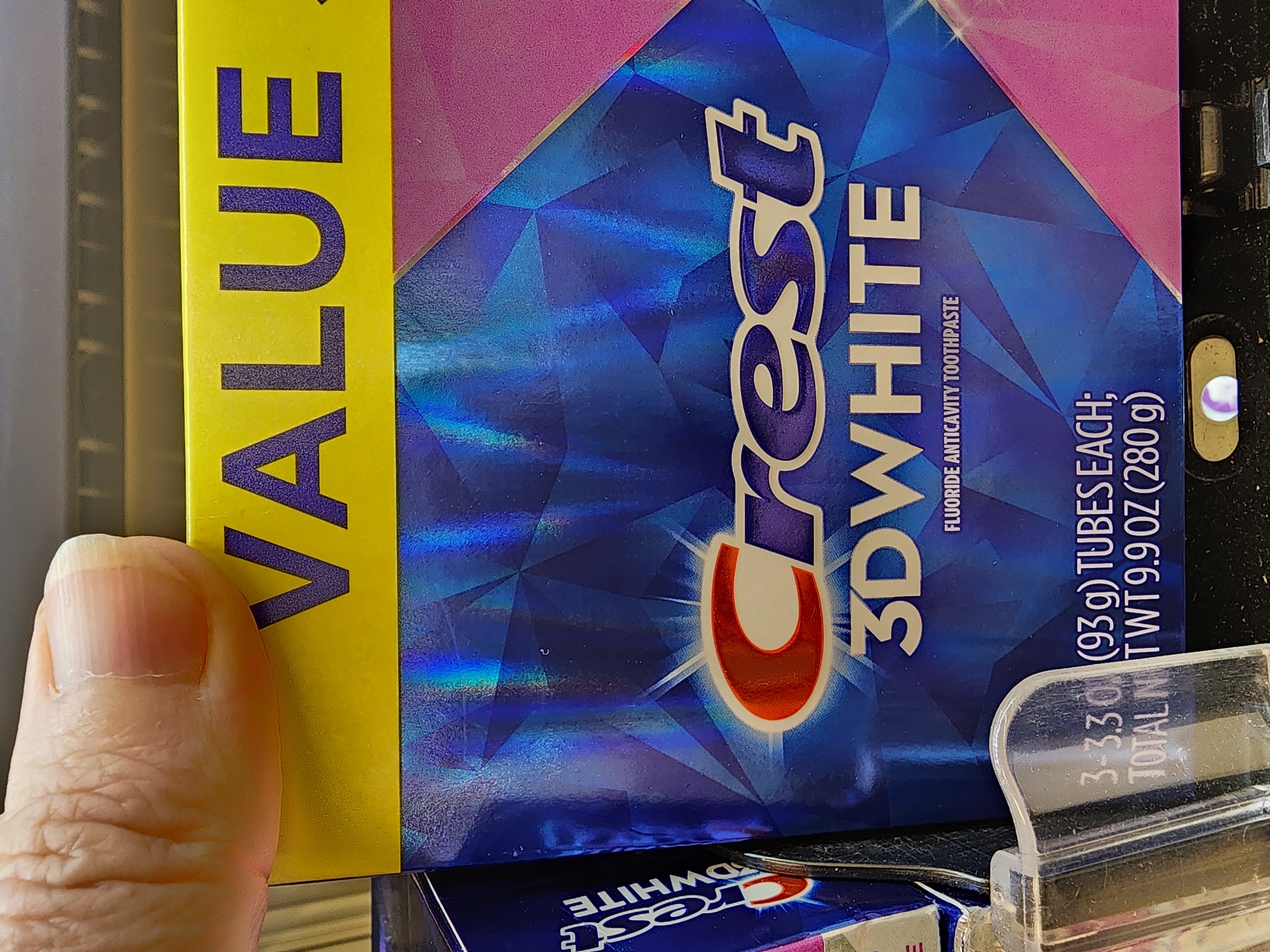 A close-up, eye-level shot shows a person's thumb and fingernail pressing down on the top edge of a yellow and blue box of Crest 3D White toothpaste. The box has a holographic, faceted design and features the Crest logo in the center. Text on the box includes "VALUE," "3DWHITE," and "FLUORIDE ANTICAVITY TOOTHPASTE." Below this, there is text indicating "3-3.3 OZ (93 g) TUBES EACH; TOTAL NET WT 9.9 OZ (280 g)." A transparent plastic cap or dispenser is visible in the lower left corner of the image. The background is dark and indistinct, suggesting an indoor setting, possibly a retail environment. The lighting is somewhat uneven, with reflections visible on the toothpaste box. The overall scene appears to be a product display. The image was captured in Camp Hill, United States of America.