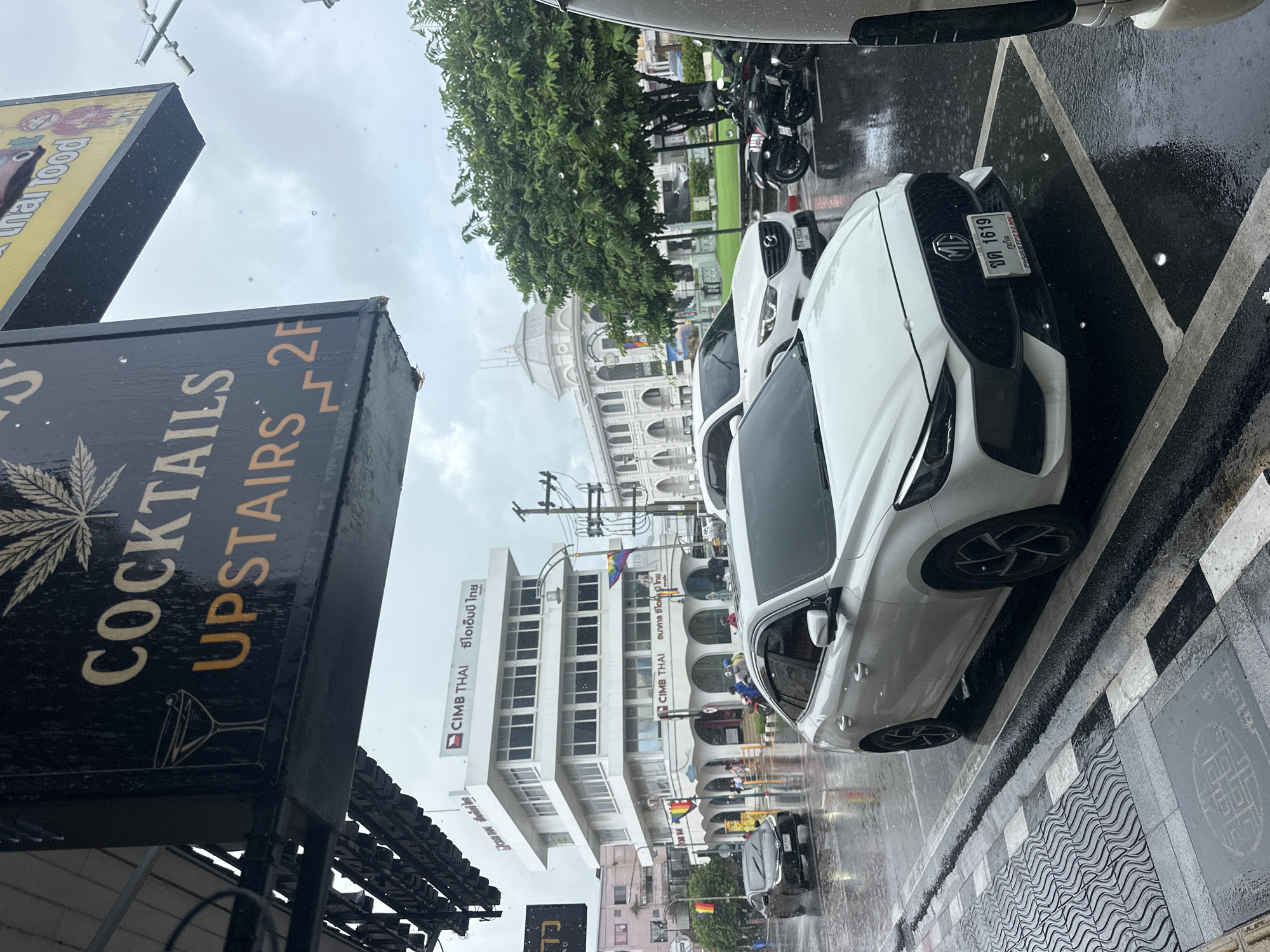 This media file depicts a street scene in Phuket City Municipality, Thailand, on a rainy day. The foreground features a dark sign with text and an illustration of a martini glass, partially obscured by water droplets. The text on the sign reads "COCKTAILS UPSTAIRS 2F".
Further into the scene, several vehicles are visible, including a white MG car with a license plate that reads "ขต 1619 ภูเก็ต PHUKETPIYA MG". The street is wet and reflective, indicating recent rainfall, with visible water on the pavement and the vehicles. In the background, a white building with arched windows and a domed structure is prominent, along with other buildings, including one with "CIMB THAI" signage. The sky is overcast and appears to be daytime. There are also motorcycles parked on the side of the road. Rainbow flags are visible on some of the buildings, suggesting a festive or inclusive atmosphere. The overall mood is that of a wet, urban environment.