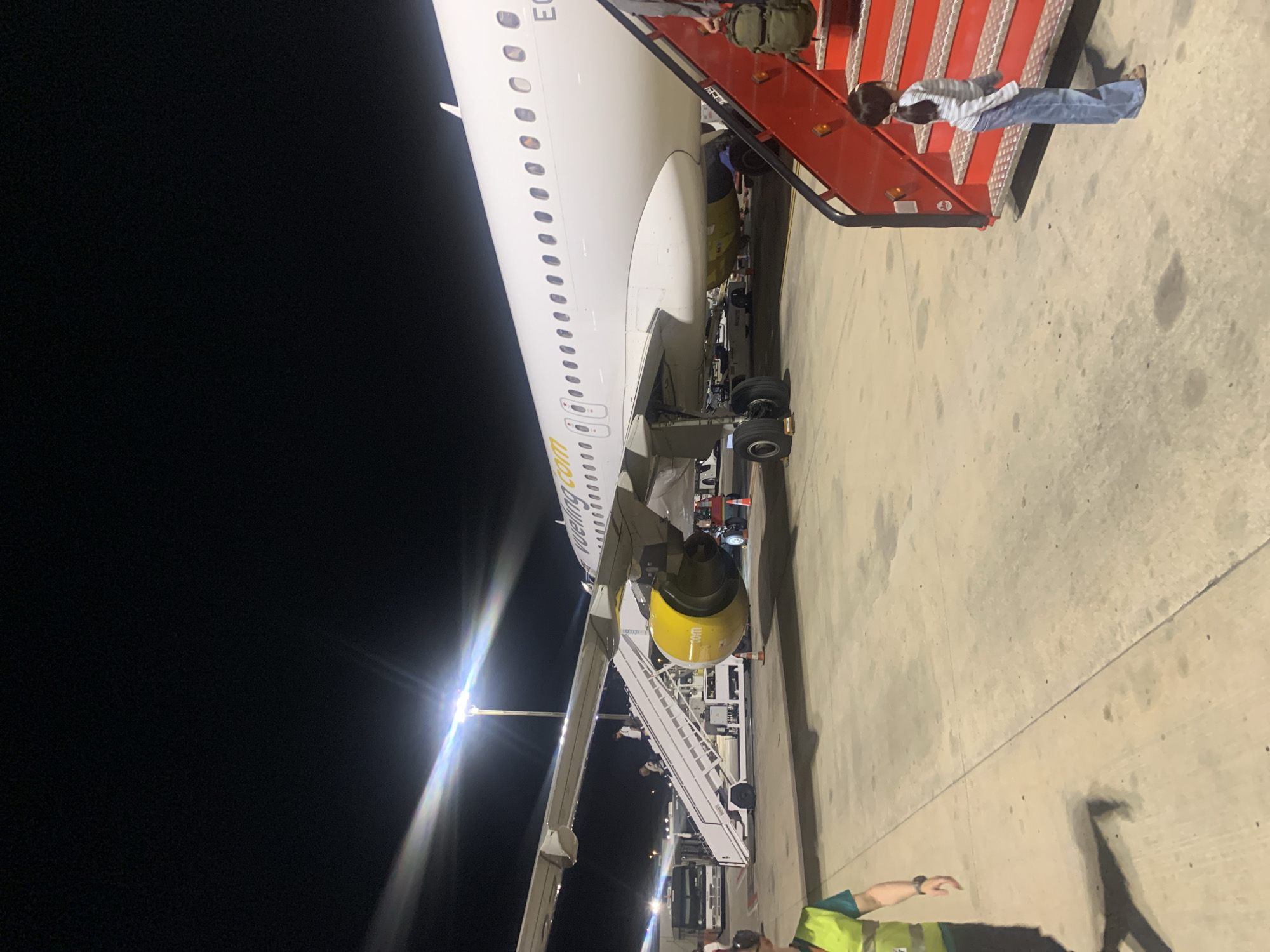 The image depicts an airport tarmac at night, likely at Barcelona–El Prat Airport (BCN) in Sant Boi de Llobregat, Spain, due to the presence of Vueling aircraft, a Spanish low-cost airline headquartered at this airport. A white Vueling airplane is parked on the tarmac, with its wing and engines clearly visible. A mobile airstair is positioned next to the aircraft, with several people ascending and descending it. A young girl in a striped shirt and jeans stands at the bottom of a red mobile boarding stairs, looking up. To the left, a person wearing a bright yellow safety vest stands on the tarmac, appearing to be airport personnel. In the background, a bus and other airport equipment are visible under the artificial lighting of the tarmac. The scene is illuminated by bright floodlights, casting long shadows on the concrete ground.