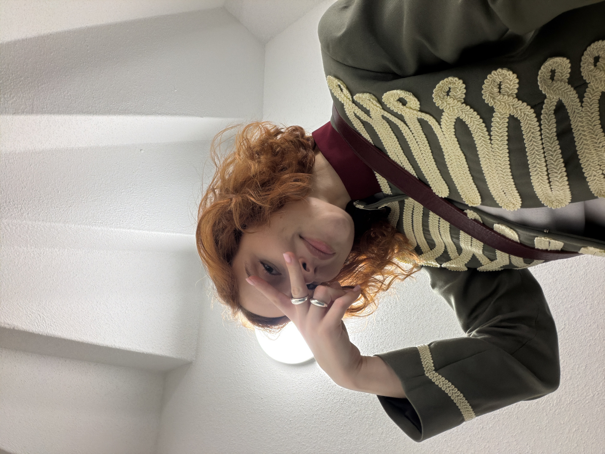 A young woman with curly, red hair and fair skin is captured from a low angle in a selfie, holding her right hand up to her face in a peace sign. She is wearing a dark green, military-inspired jacket with ornate gold braiding across the chest and sleeves, a maroon collar, and a maroon strap across her chest. She has two silver rings on her right hand. The background consists of a brightly lit white wall with geometric shadows cast by architectural features, suggesting an indoor stairwell or hallway. The location is not identifiable as Strasbourg, France.