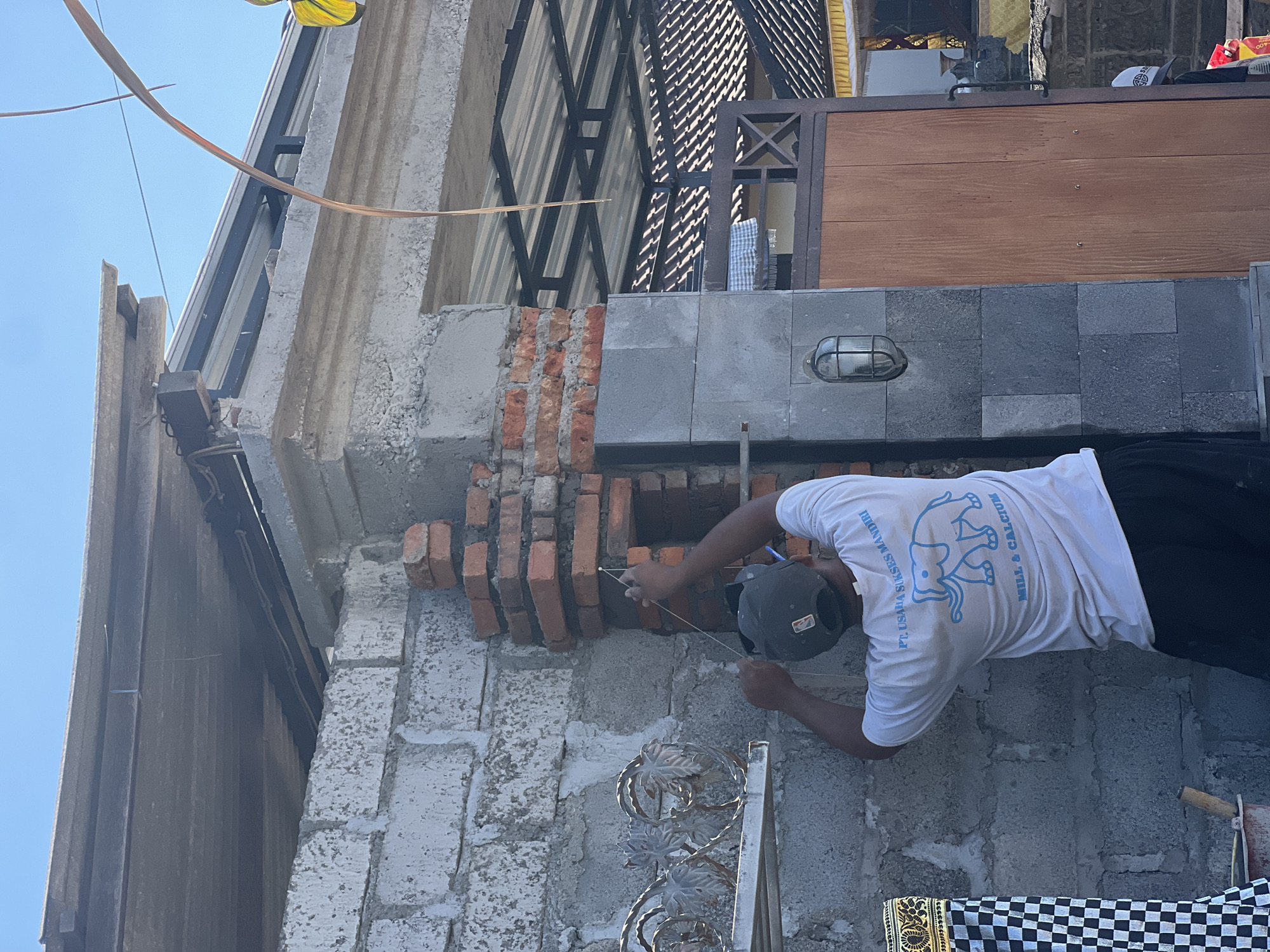 A person, wearing a white t-shirt with a blue elephant graphic and text, and a dark baseball cap, is performing masonry work on a wall. The individual is laying bricks, using string to guide the placement. The scene appears to be an outdoor construction site, likely in Sukawati, Indonesia, given the context. The wall is made of concrete blocks, and some sections are already plastered. There are also exposed red bricks in various stages of being laid or dismantled. The background shows elements of a building, including a corrugated metal roof with a lattice structure, and a wooden gate. A light fixture is visible on the gray stone portion of the wall. The lighting suggests it is daytime, with bright sunlight casting shadows. The person's focused posture indicates an active work environment. Visible text on the t-shirt reads "PT USAHA SUKSES MANDIRI" and "MILL & CALCIUM".