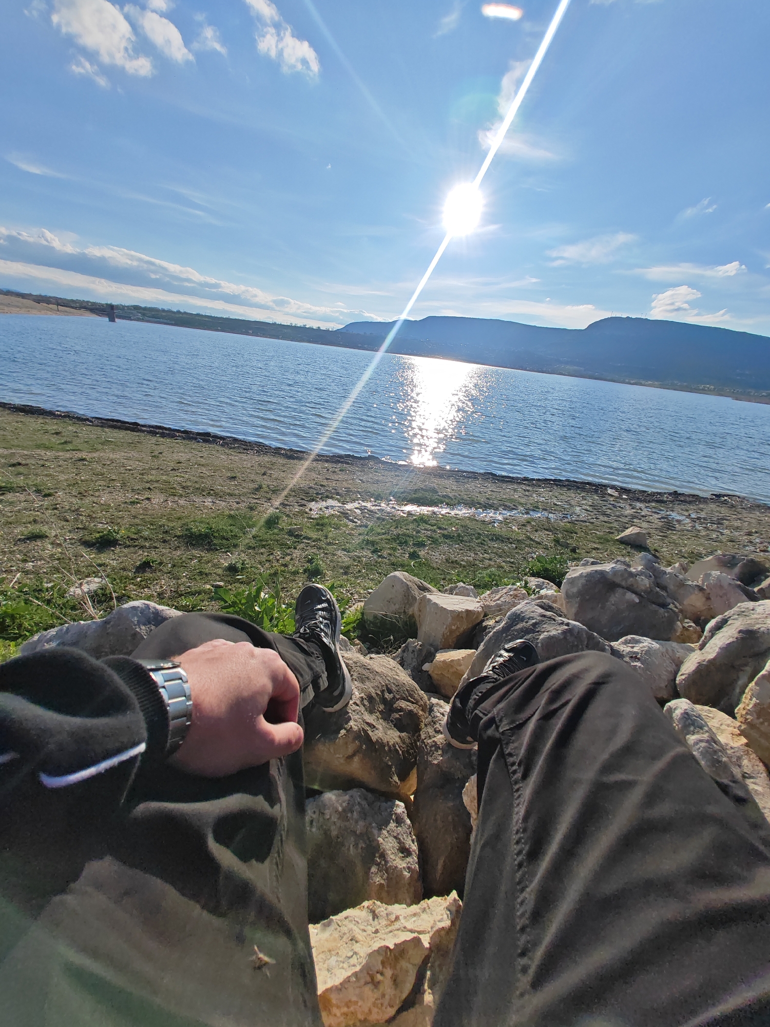 A medium shot captures the first-person perspective of a person sitting among rocks near a body of water. The individual is wearing black pants, a black jacket with white trim, a silver watch, and black sneakers. Their left hand rests on their left leg, which is propped up on the rocks. The right leg is also extended forward, resting on the rocks.
In the background is a body of water that reflects sunlight, with land visible across the water. The sky is blue with scattered white clouds and a prominent sun glare. The foreground also includes some grass and dirt at the water's edge, transitioning to the rocky area where the person is sitting.
The photograph was taken near the "Kyoshkovete" area of Shumen, Bulgaria, known for its dam and reservoir used for irrigation and recreation.