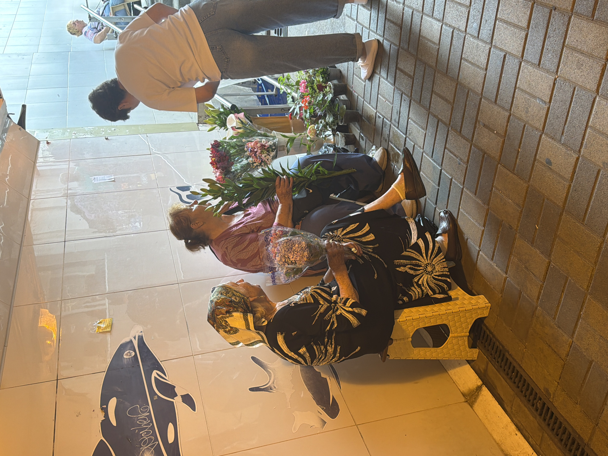 The image captures a street scene in Sochi, Russia, with three individuals in the foreground. On the left, an elderly woman wearing a headscarf and a black and white patterned dress is seated on a yellow stool, holding a bouquet of flowers. To her right, another woman, also seated, in a pink top and dark pants, is holding a larger bouquet of flowers. A third person, viewed from the back, is standing and appears to be interacting with the seated women, possibly as a customer. They are dressed in a light-colored t-shirt and blue jeans. In front of the standing person and between the seated individuals, there are potted plants and more flowers displayed. The background features a tiled wall with a blue mural of fish and a metal structure above. The ground is paved with brickwork. The overall lighting suggests it is either early morning or late evening, casting warm tones on the scene.