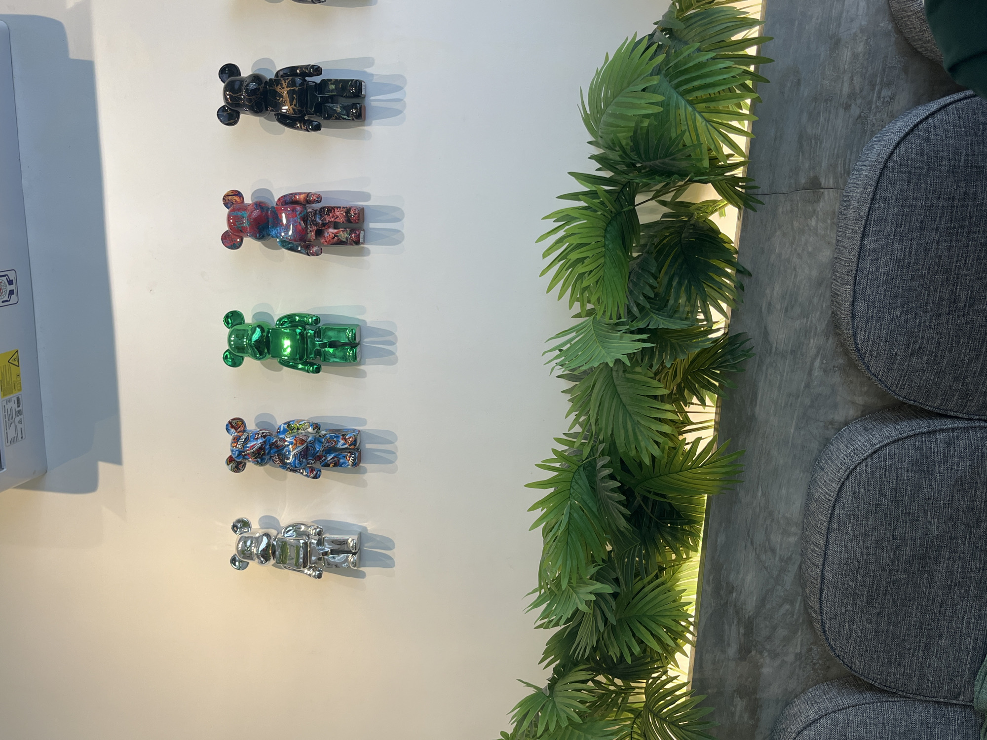 A row of six decorative bear figurines is mounted on a white wall in a room. From left to right, the figurines are silver, blue with a colorful pattern, green, red with a red and blue pattern, black with gold accents, and partially visible black with gold accents. They are positioned above a decorative arrangement of artificial green palm fronds, which are illuminated from behind by a warm light strip. Below the palm fronds is a concrete wall section, and at the very bottom of the image, the tops of three dark gray upholstered chairs are visible. The scene appears to be indoors, possibly in a commercial establishment like a cafe or shop, given the decorative elements. This scene is located in Kuta Selatan, Indonesia.