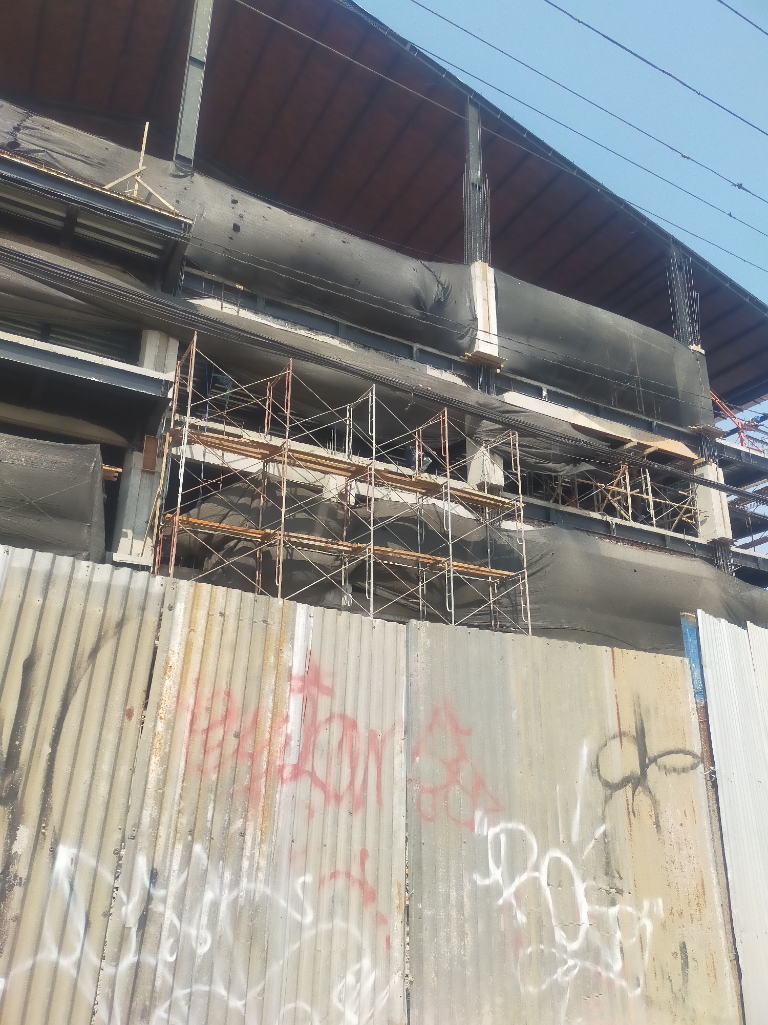 The image shows a construction site in Talang, Indonesia. A multi-story building is under construction, with scaffolding visible on the exterior. The building is partially covered with black netting. A corrugated metal fence, covered in graffiti, stands in the foreground, separating the viewer from the construction site. The sky is clear and blue, with power lines running across the upper portion of the image. No people are visible in the image.