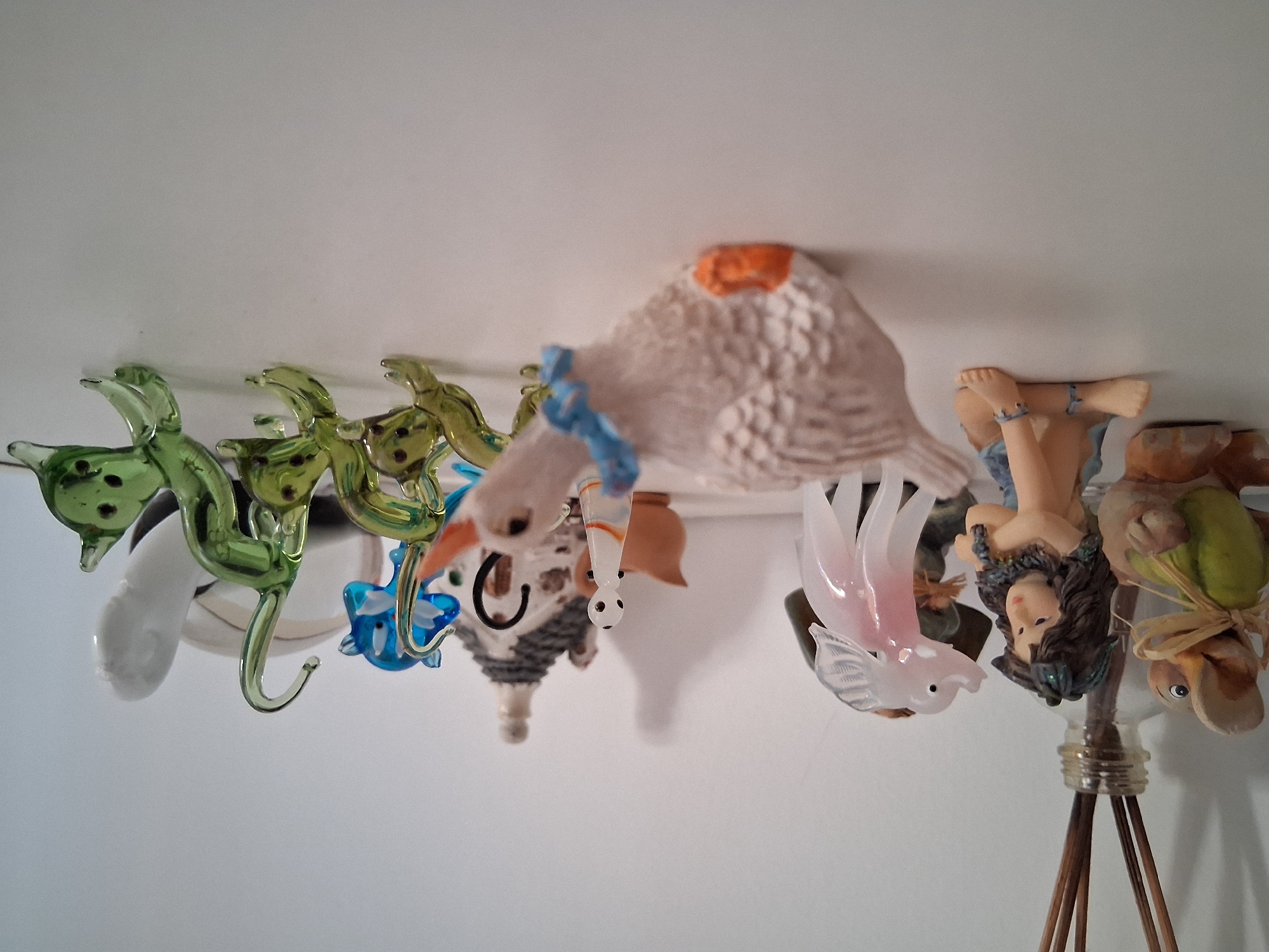 The image shows a collection of decorative objects hanging from a white ceiling, possibly in a home or a shop in Colorno, Italy. The objects are diverse and appear to be made of glass, ceramic, and other materials.
On the left, there are several green glass objects that resemble leaves or abstract shapes, some with dark spots. Further to the right, there is a white object that could be a stylized bird or a cloud. In the center, a large, textured object that looks like a white bird with an orange beak or cap dominates the scene. Below this bird, there's a smaller, more detailed ceramic figure that appears to be a house with a chimney and a small character with black dots for eyes hanging from it.
To the right of the central bird, there are more whimsical figures. A translucent pink glass bird with detailed wings is prominent. Next to it, a figurine of a woman with dark hair, wearing a blue necklace and a tan skirt, is suspended upside down. To her right, another upside-down figurine appears to be a duck or a similar bird, wearing a green garment and tied with a straw ribbon. These figures are hanging from a string or wire attached to a glass bottle at the bottom right, which has some wooden sticks emerging from it.
The lighting suggests it might be daytime, with the light source coming from above, casting subtle shadows on the white surface. The overall impression is one of a quirky and eclectic collection of decorative items. No text is visible in the image.