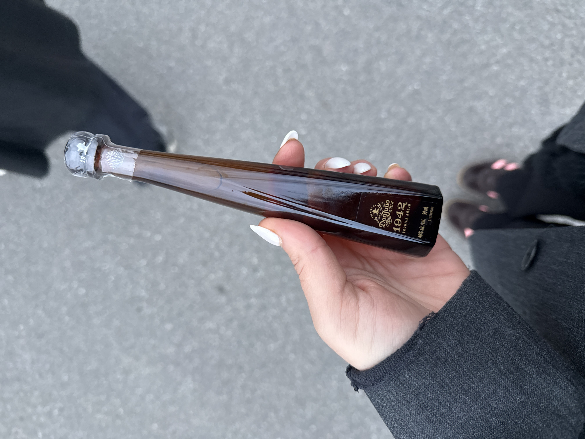A hand with long, white fingernails holds a small bottle of Don Julio 1942 tequila against a gray asphalt background. The bottle is amber-colored with a clear glass stopper and is partially obscured by the hand. The background shows blurred figures and feet in the distance. The location is not specified as Croix, France, in the image. The attire includes a dark gray jacket.