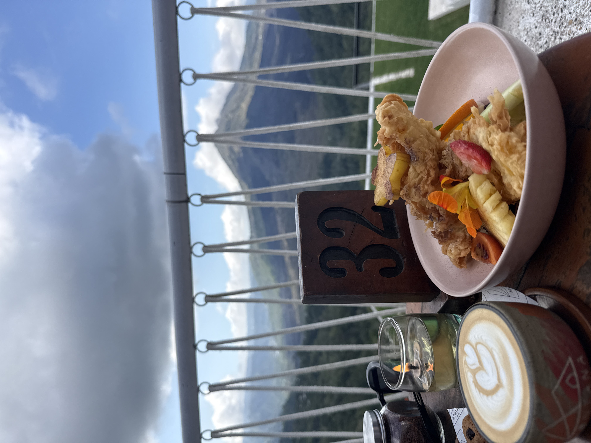 This is a close-up shot of a table at a restaurant in Kintamani, Indonesia, with a scenic mountain view. The table is set with a coffee cup featuring latte art, a small candle in a glass of oil, a jar of coffee grounds, a wooden table number that reads "32," and a pink bowl filled with food. The food appears to be a fried item, possibly chicken or fish, accompanied by sliced fruits like pineapple, strawberry, and orange, and some green vegetables.
In the background, a railing is visible, beyond which lies a breathtaking landscape. Rolling hills covered in lush greenery stretch into the distance, culminating in a majestic mountain, likely Mount Batur, dominating the horizon under a cloudy sky. The overall impression is one of a relaxed dining experience with a stunning natural backdrop, typical of Kintamani's popular viewpoints.