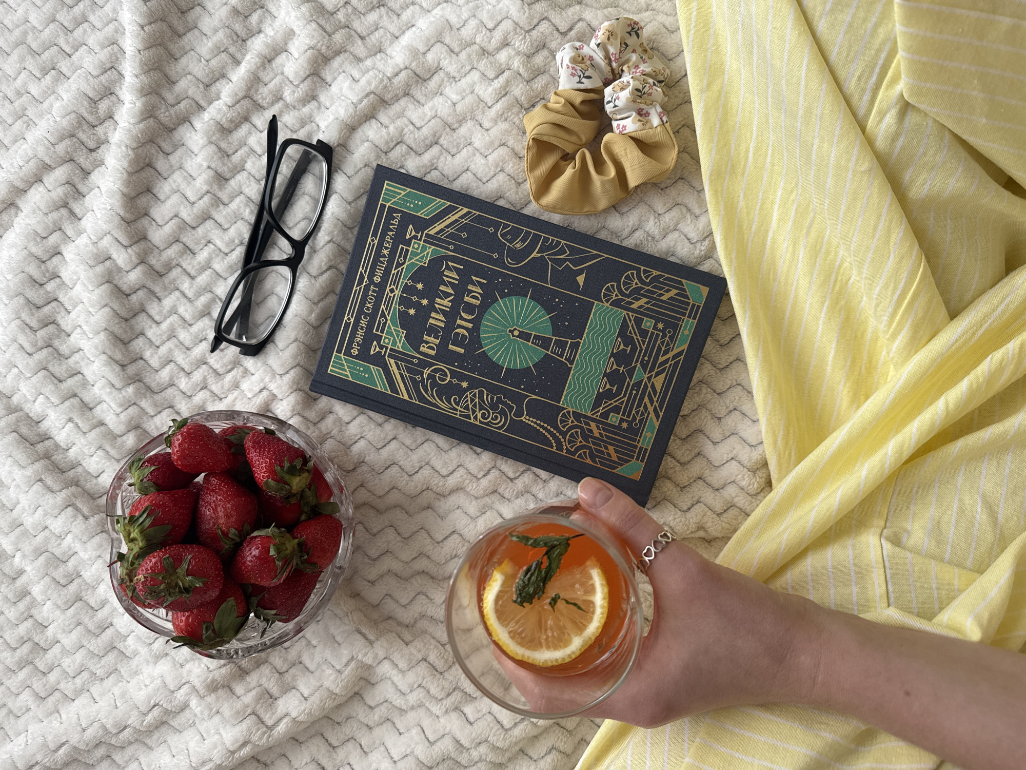 The image shows a flat lay arrangement on a white textured blanket. A pair of black eyeglasses and a bowl of strawberries are placed on the left side. A book with a navy blue cover and gold designs, along with two scrunchies, are arranged in the upper right quadrant. A hand holds a glass with a beverage containing a slice of lemon and mint leaves in the lower right corner. The image suggests a casual, relaxed setting. The text on the book cover is in Cyrillic. There is no indication in the image of a location in Izhevsk, Russia.