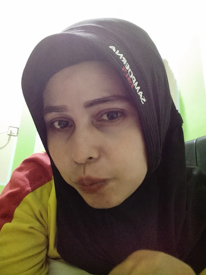 The image depicts a female individual, likely an adult, in a headshot taken indoors in Ranugedang, Indonesia. She is positioned slightly to the right of the frame, looking directly towards the viewer with a neutral expression and slightly pursed lips. Her skin tone is light to medium, and she has dark eyes with defined eyebrows. She wears a black hijab that covers her head and neck. Reversed white and red text, likely "SAMPOERNA," is visible on the upper left side of her hijab. Beneath the hijab, a garment with distinct red and yellow sections is visible on her left shoulder and arm. The background consists of an interior wall, with a light green section on the left and a white section above and to the upper left. A white electrical outlet is mounted on the light green wall. A dark, indistinct object, possibly part of a chair, is partially visible on the right side of the frame.