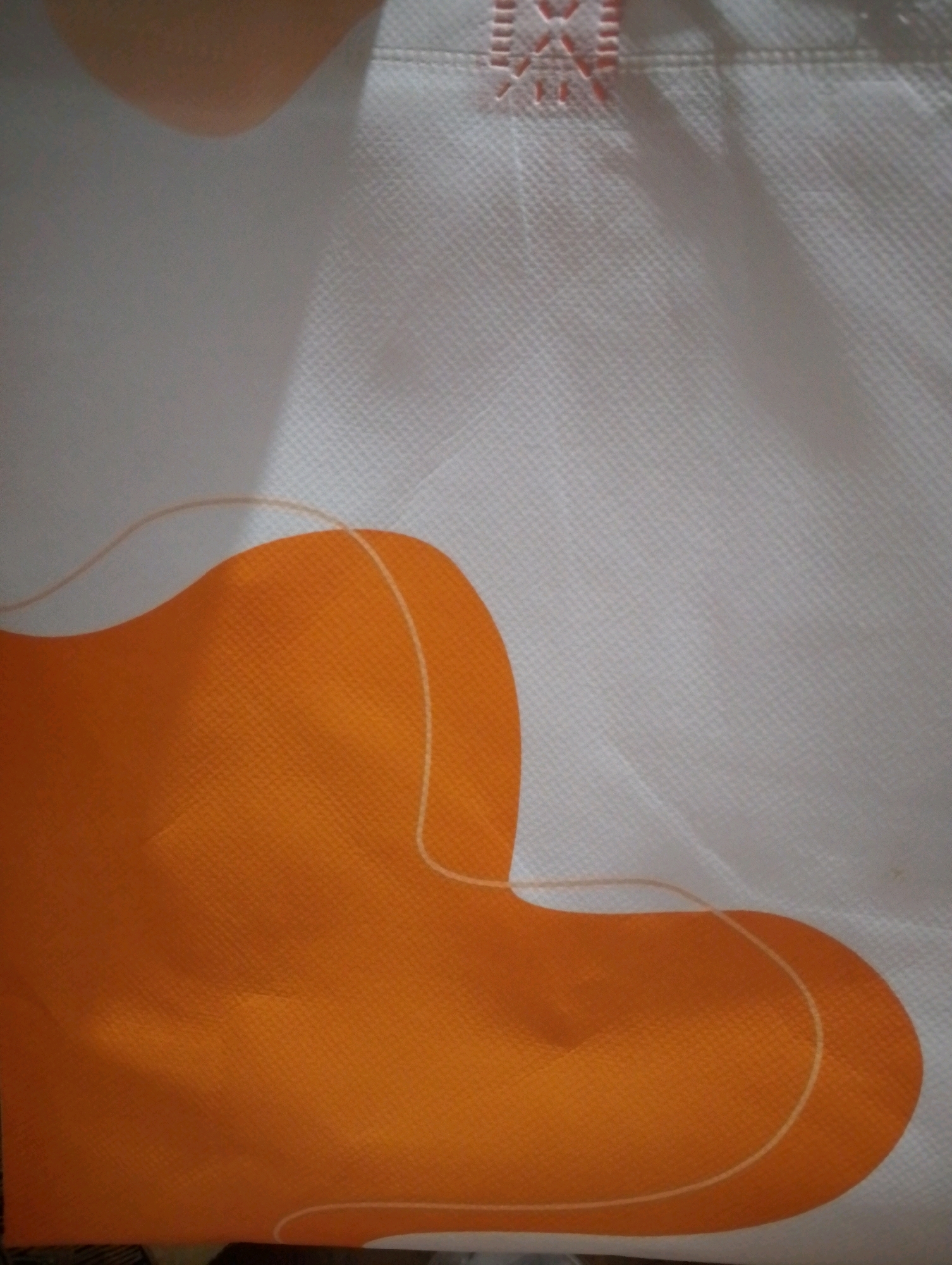 An abstract image displays a white fabric surface with an orange blob-like design. The orange design has a thin, light-colored outline that follows its shape. A hint of a human hand is visible in the upper left corner, casting a shadow. The fabric has a textured appearance with a grid-like pattern.