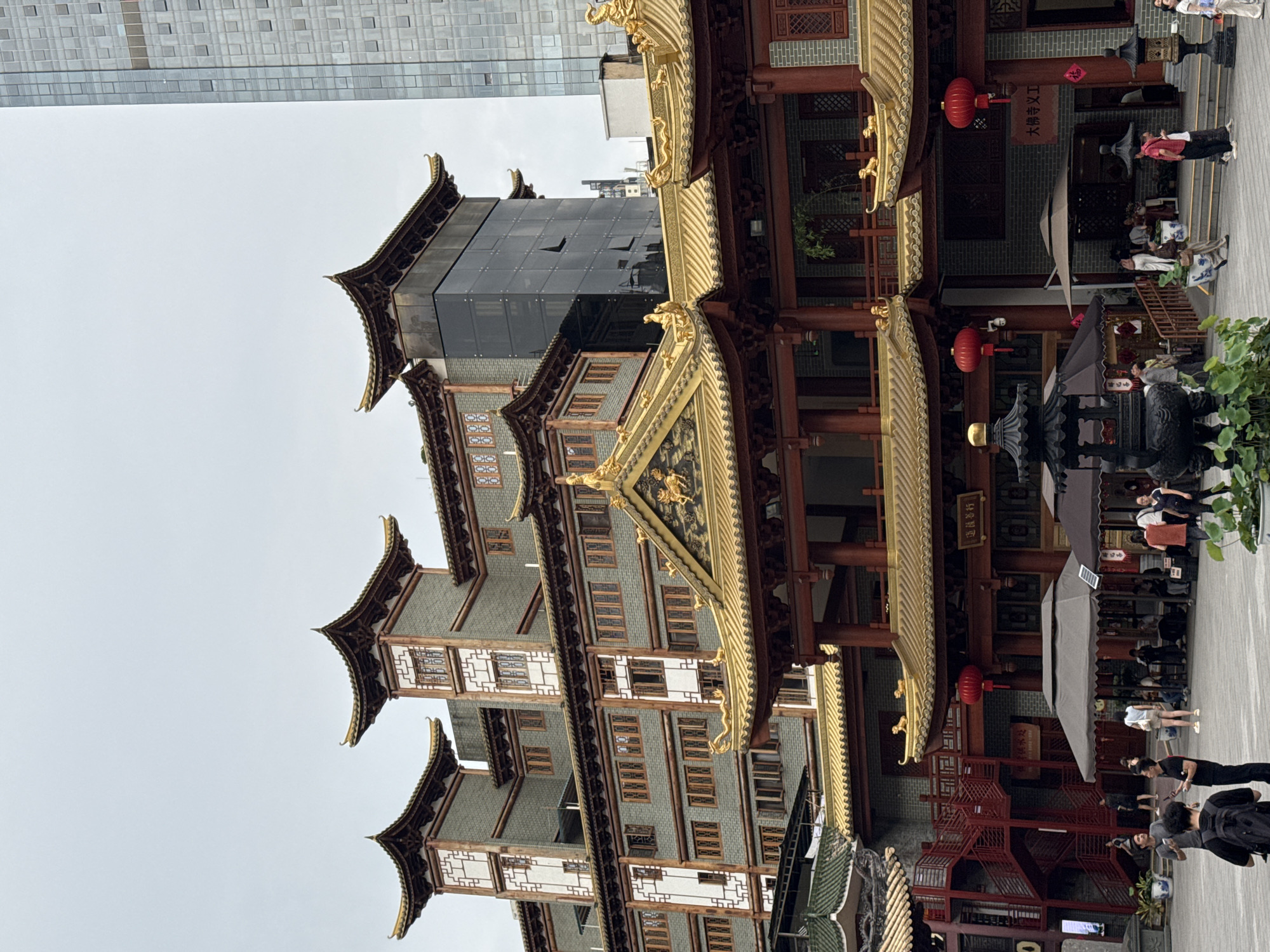 The image captures a bustling scene in the Yuexiu District of China, dominated by a traditional multi-story Chinese building featuring a distinct pagoda-style roof, gold embellishments, and intricate window patterns. Adjacent to this historical structure is a modern building with a glass facade, highlighting a contrast between architectural styles.
Numerous people are present, walking or standing within the building's courtyard and surrounding areas, suggesting a public or commercial space. Red lanterns hang from the building, adding to the cultural atmosphere.
The environment appears to be urban, with overcast skies providing soft lighting. The scene suggests a blend of historical preservation and contemporary urban development.
