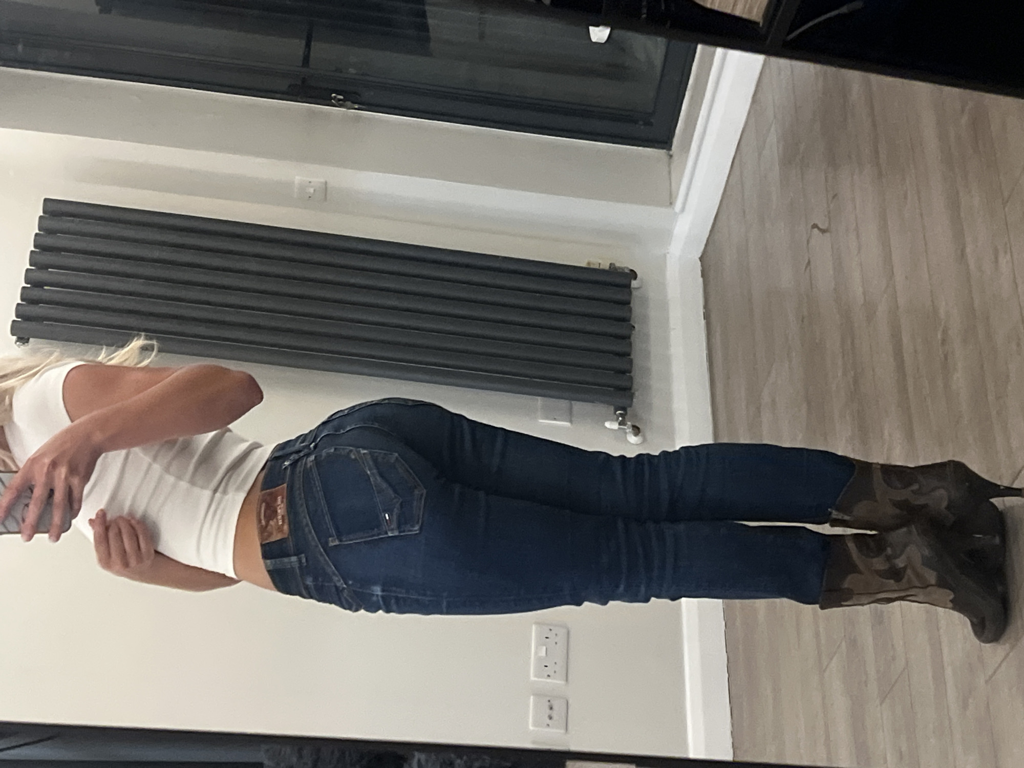 A woman is standing indoors, likely in a residential space, and is captured in a mirror. She is wearing a white tank top, fitted blue jeans, and high-heeled camouflage boots. The background includes a grey horizontal radiator attached to a light-colored wall, and electrical outlets are visible. The floor is light wood. The scene appears to be in a modern, tidy environment. No specific location in London is identifiable from this image.