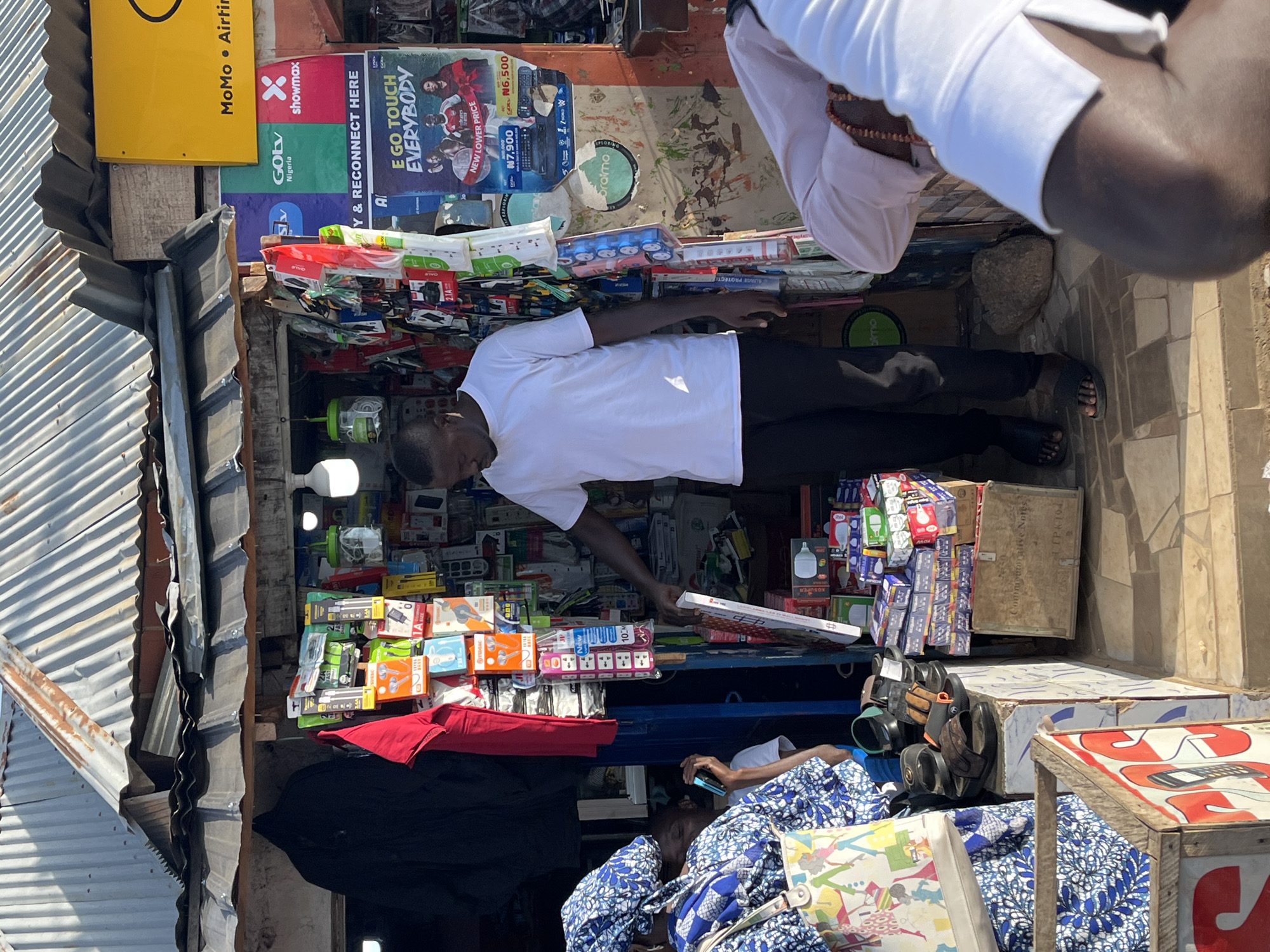 The image depicts a bustling street-side stall in Gura Top, Nigeria, likely a mobile phone accessory shop. A man in a white t-shirt and black trousers stands behind the counter, actively engaged with customers. He is holding a white box, possibly for a product. To the left, a woman dressed in a vibrant blue and white patterned outfit, including a headscarf, is browsing. Another person, wearing a light pink shirt and a beaded necklace, is visible to the right, seemingly interacting with the stall.
The stall itself is densely packed with various items. Shelves are overflowing with packaged mobile phone accessories like earphones, chargers, and SIM card packaging, indicated by colorful branding and text. Various posters advertising services and products are plastered on the back wall, including logos for "MoMo" and "Showmax," suggesting telecommunication and entertainment services. A bright yellow sign is also visible at the top right.
The stall has a corrugated metal roof, typical of many informal businesses in the region. Various footwear, including sandals, are stacked near the entrance, suggesting a diverse range of goods are sold. The ground is paved with tiles, and a rough stone lies on the ground near the man. The overall impression is one of a lively, commercial environment.