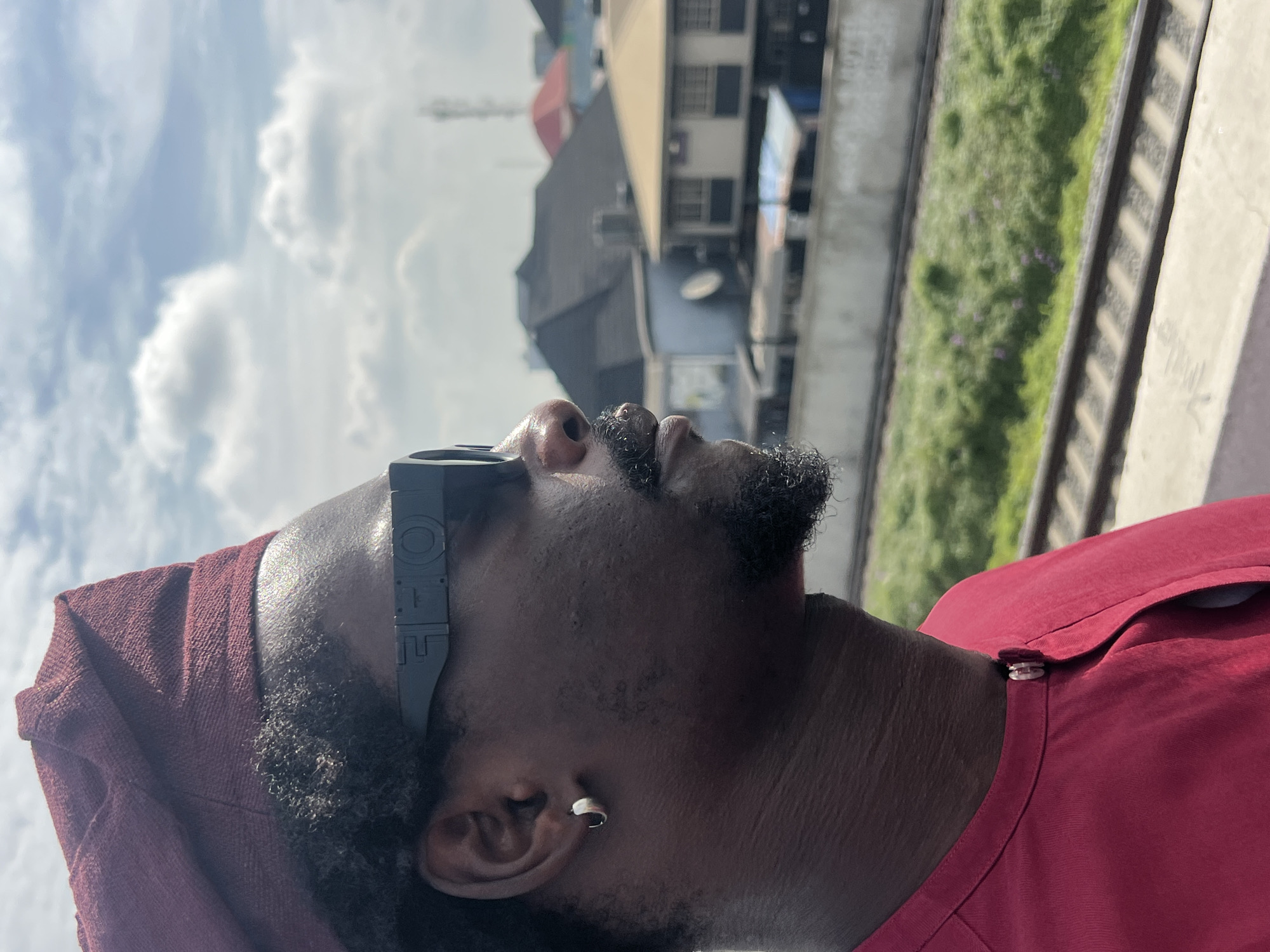 This vertically oriented image captures the profile of an adult male looking upwards towards a cloudy sky. The subject has dark skin, a neatly trimmed beard, and is wearing a maroon-colored shirt with a matching head covering or cap. A small silver hoop earring is visible in his left ear. He is wearing a unique pair of black glasses or goggles, with the text "IOE" visible on the side temple.
The background, which is out of focus, reveals an urban or suburban setting. Several buildings are visible, including one with multiple windows and another with a dark roof. Below these structures, what appear to be train tracks or a raised concrete platform with green vegetation run parallel across the mid-ground. The sky above is bright with scattered white cumulus clouds, indicating daytime. The man's upward gaze suggests he is observing something in the sky or distance. The overall scene is consistent with an outdoor location in Ikeja, Nigeria.
