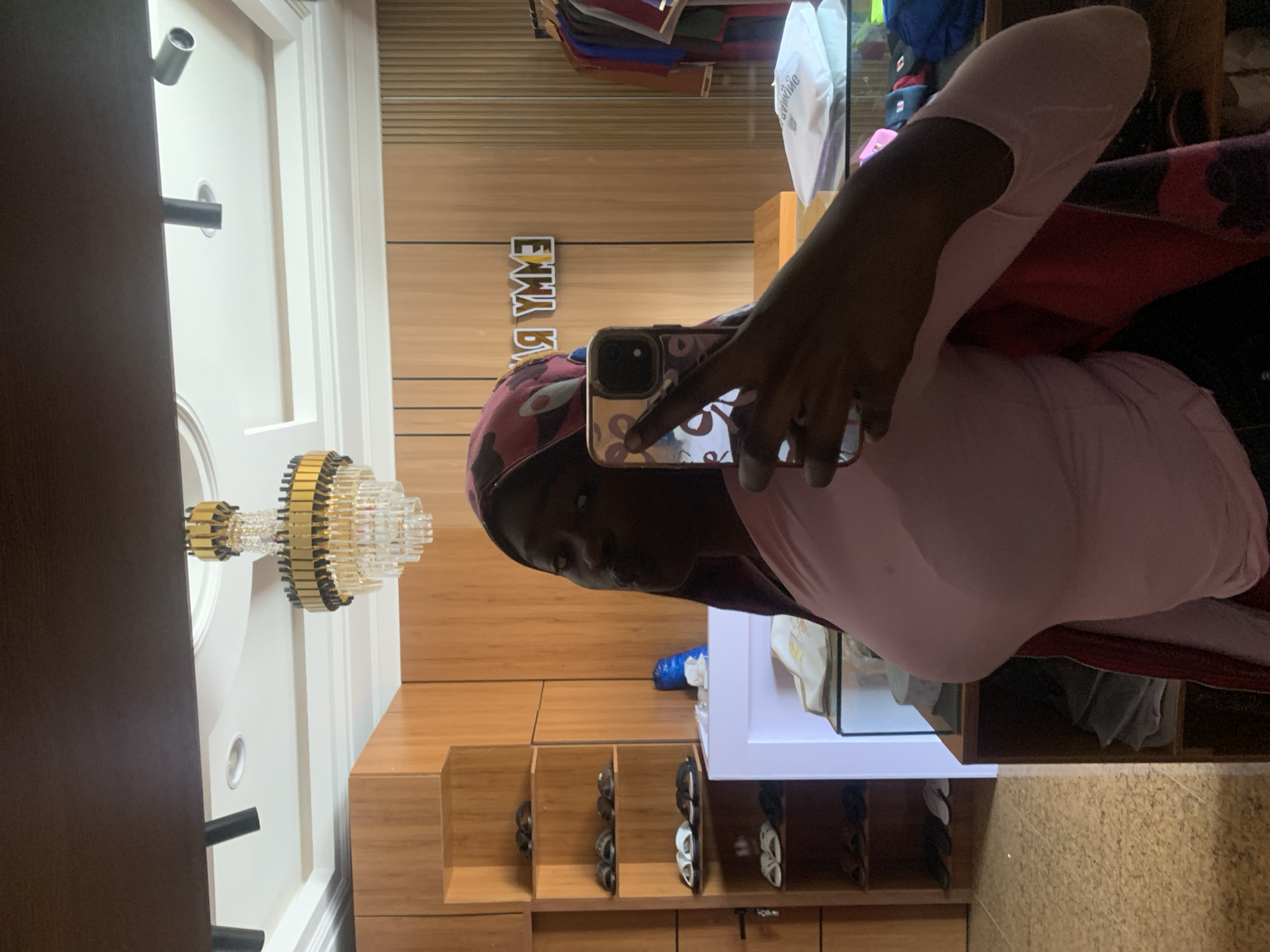 A young Black woman is taking a mirror selfie in what appears to be a retail space. She is wearing a light pink long-sleeved shirt and a patterned headscarf. The background shows a display of shoes on shelves, a white display case, wooden paneled walls, and a chandelier on the ceiling. The woman's face is partially obscured by her phone, which she is using to take the photo. There is a clothing rack with various garments visible in the reflection of the mirror. The overall lighting suggests an indoor environment, likely a boutique or shop. The location, Gura Top, Nigeria, is indicated by text on the wall in the reflection, which reads "EMMY R".