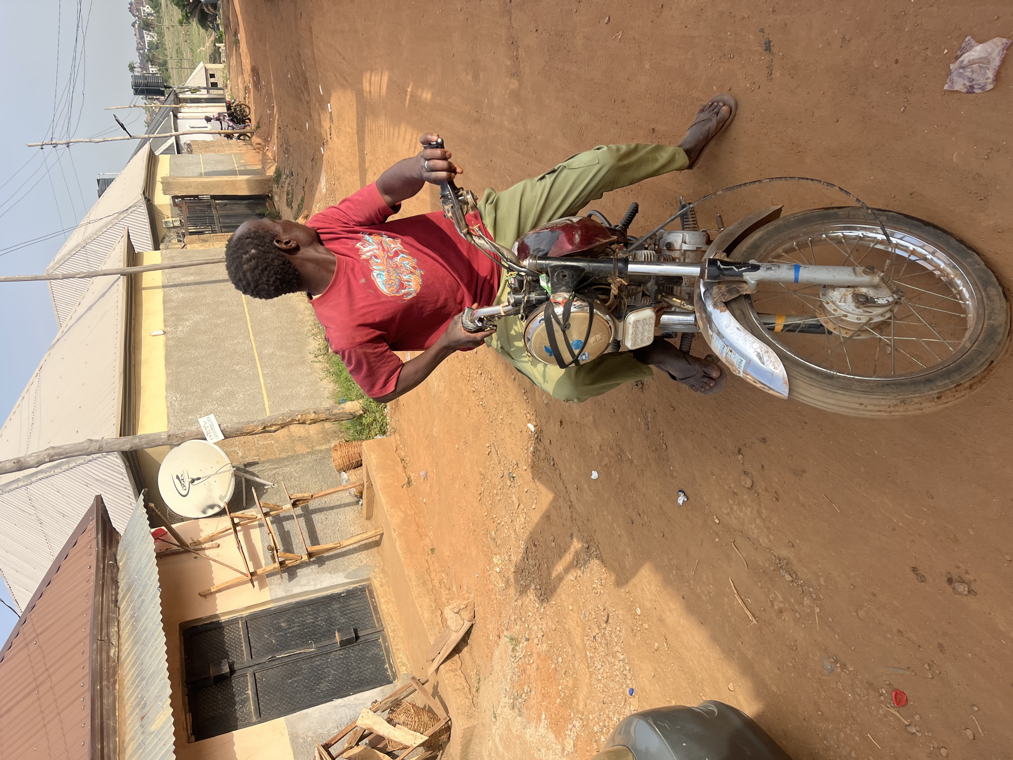 This image captures a bright, daytime scene on a dusty, reddish-brown unpaved road in what appears to be a residential or semi-urban area. The central subject is a dark-skinned man, seen from a slightly elevated perspective, seated on a motorcycle. He wears a red long-sleeved shirt with a graphic design, olive green pants, and brown sandals. His short, dark hair is visible, and he is looking towards the left side of the frame (his right). He is holding the handlebars of the motorcycle, which has a reddish-brown fuel tank, silver handlebars, front forks, and a front fender over a spoked wheel.
In the background, several single-story buildings with corrugated metal and flat roofs are visible, painted in shades of yellow, beige, and grey. Utility poles with overhead lines run through the scene. A white DSTV satellite dish is mounted on a building. Next to it, a white sign is visible, displaying black text that reads "HOUSE FOR SALE / FOR RENT / 08064441584".
The ground is dry, with pebbles and scattered pieces of litter, including a red plastic bottle cap and white plastic debris. The clear, bright sky suggests sunny weather. The overall environment, including the unpaved roads and building styles, aligns with typical scenes found in developing areas, consistent with the provided location context of Abuja, Nigeria. The man appears to be either stationary or in the process of operating his motorcycle.