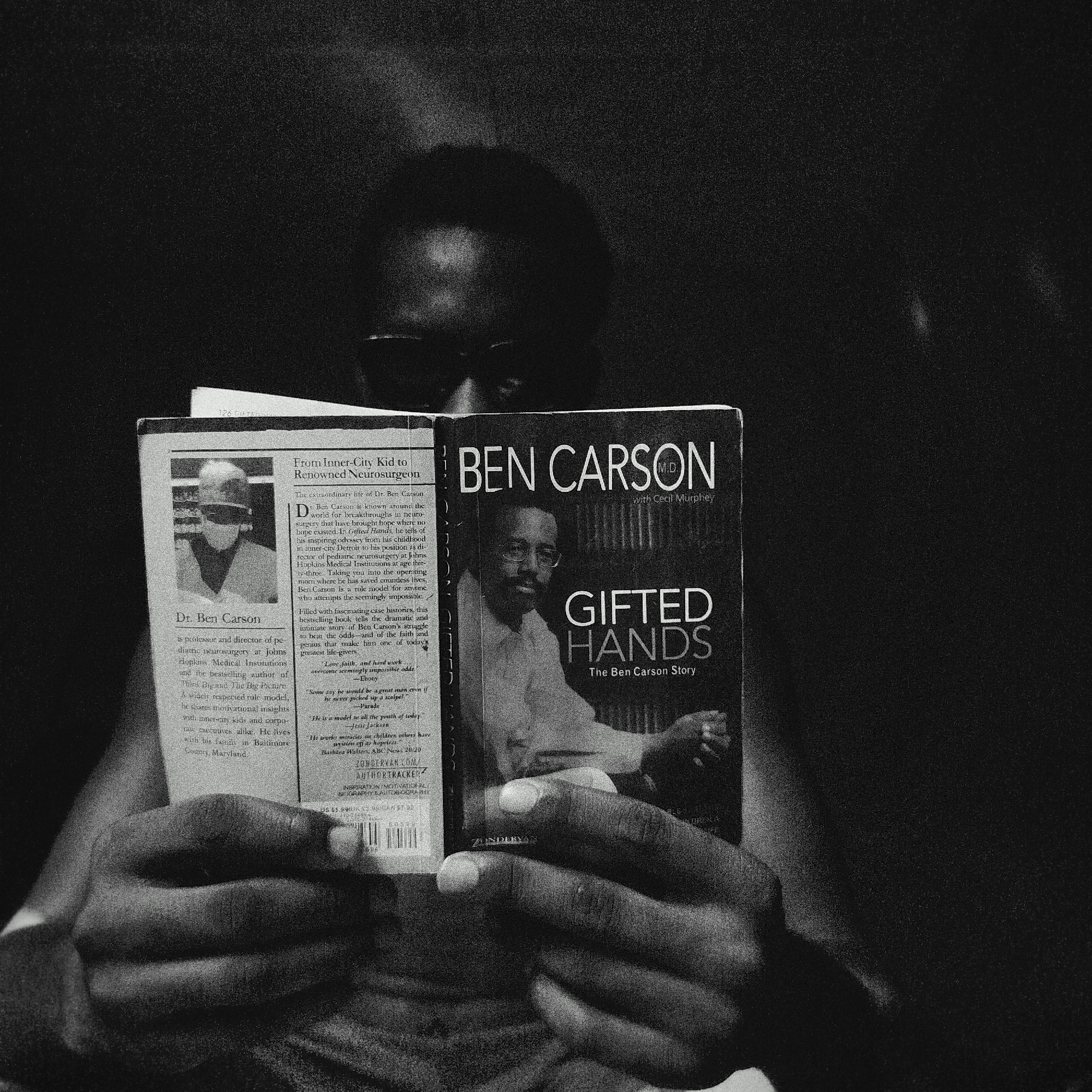 A person is reading the book "Gifted Hands" by Ben Carson. The book is open to the front cover and an inside page that contains a photo of Dr. Ben Carson and text that includes the blurb "From Inner-City Kid to Renowned Neurosurgeon." The person's face is partially visible above the book, appearing to wear glasses. The scene is black and white and grainy. There is no explicit reference to the location being Keffi, Nigeria.
