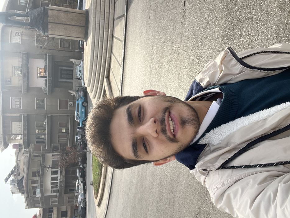 A young man takes a selfie outdoors in what appears to be Al-Muhajerin Municipality, Syria, during a bright daytime. He looks at the camera with a slight smile, having dark hair and a neatly trimmed beard. He is dressed in a beige jacket featuring a furry white lining, worn over a dark blue sweater and a white collared shirt.
The background reveals an urban setting. To the left and behind the man, several multi-story buildings with windows and balconies are visible, characteristic of residential or mixed-use architecture, with some air conditioning units attached to their facades. In the mid-ground, a prominent, round, tiered stone structure, possibly a monument base, stands with a dark-colored statue or sculpture on top. A paved street or plaza occupies the foreground and mid-ground, where several cars are parked, including a distinct light blue car. Patches of green landscaping are also present. The scene suggests a casual moment on a clear day in a bustling city environment. No discernible text is visible.