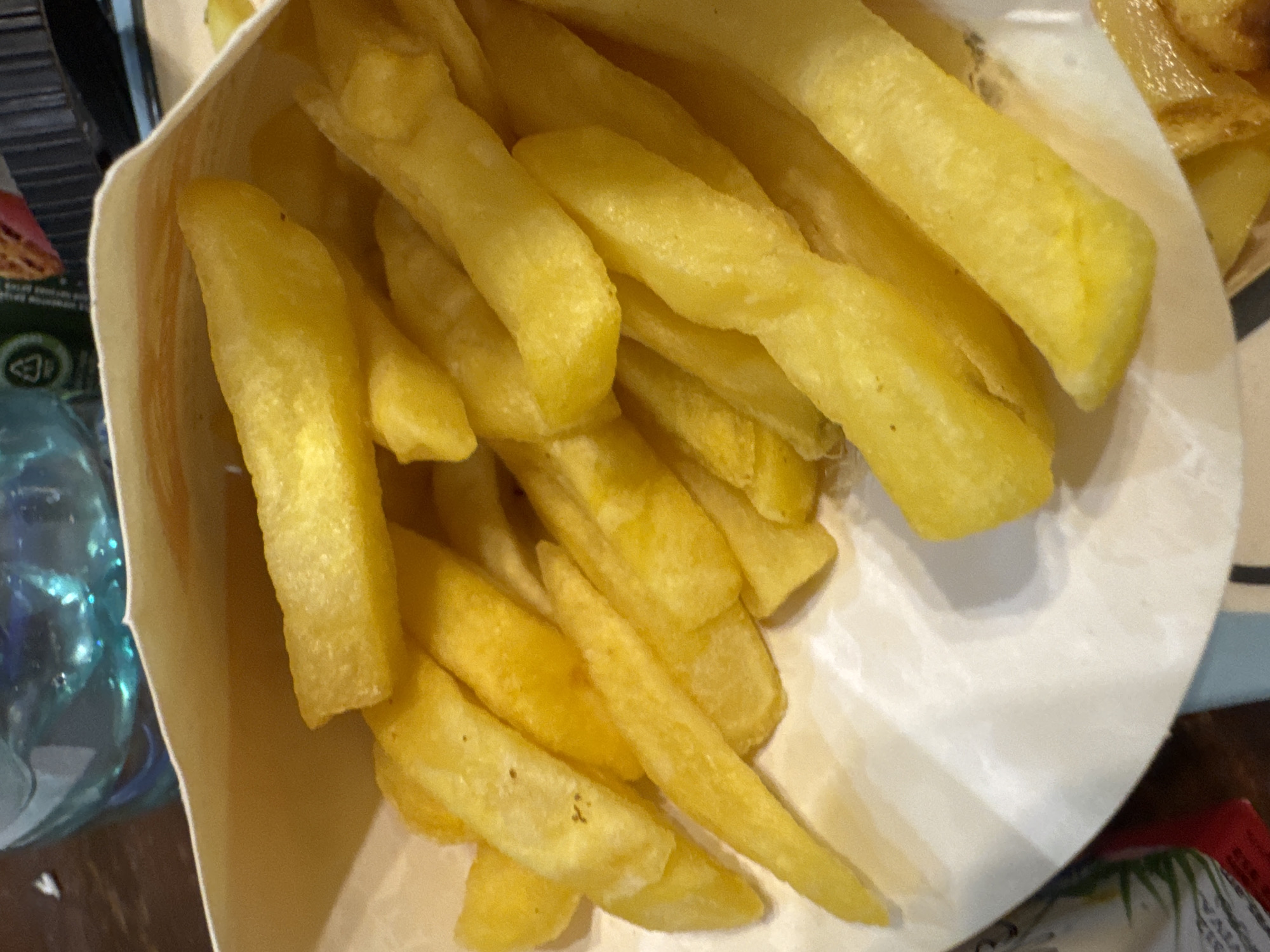 Close-up shot of a paper container filled with freshly fried French fries. The fries are golden yellow in color, and varying in size and shape. A partially visible water bottle and a portion of a food wrapper are located next to the container, sitting atop a dark wooden surface. The image was taken indoors under artificial lighting. The location of the photograph is Yerevan, Armenia.