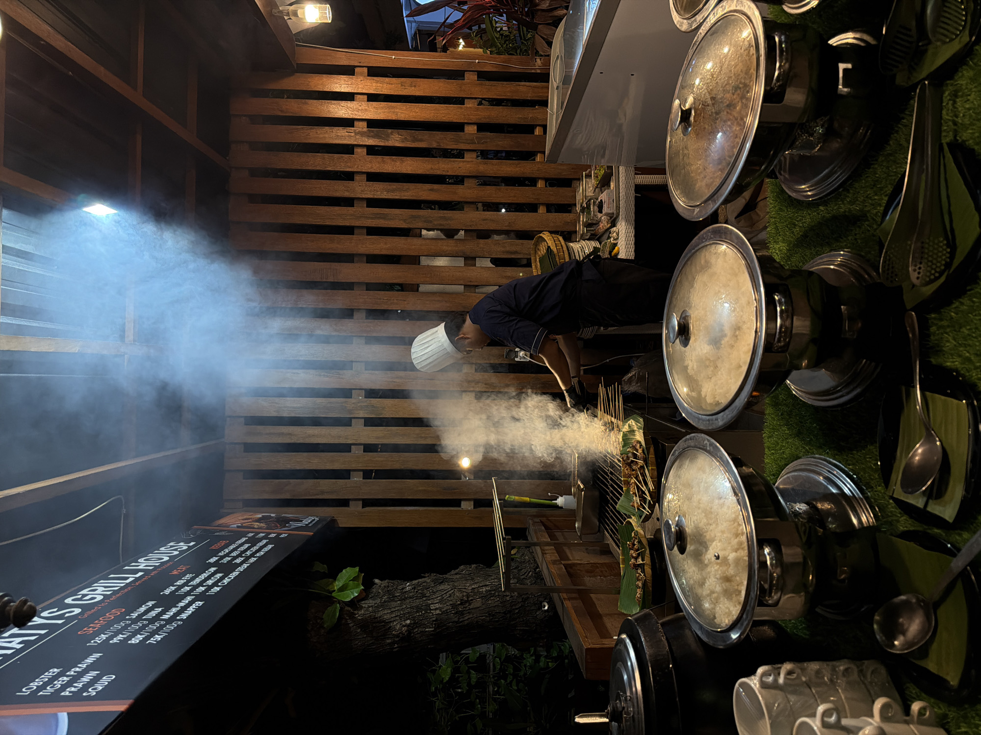 In Unknown, Indonesia, a chef wearing a white toque and dark uniform is grilling skewers of meat over an open flame. Smoke billows from the grill, obscuring the chef's face and adding a hazy atmosphere to the scene. In the foreground, a buffet-style spread of food is displayed, featuring large metal chafing dishes with glass lids, one filled with white rice and another with a darker, possibly stewed dish. A stack of white mugs and several serving spoons rest on black plates lined with banana leaves. The buffet is set on a green, grass-like mat. Behind the grill, a wooden slatted wall provides a backdrop. A dark tree with visible roots is to the left of the frame, hinting at an outdoor or semi-outdoor setting. A menu board hanging above the grill lists various seafood and meat options with prices. The lighting suggests it is nighttime or a dimly lit interior space, with a single light fixture visible above the wooden wall.