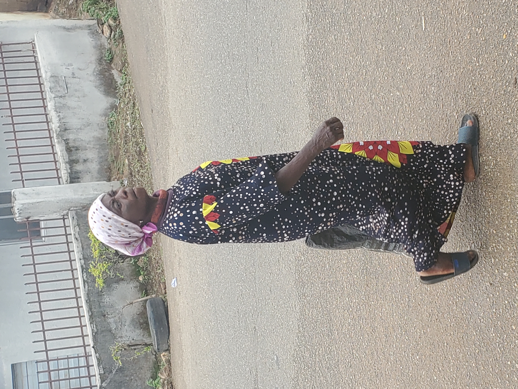 A full-length shot shows an older woman walking on a paved road. She is wearing a black and white polka dot dress with red and yellow floral accents around the shoulders and hem. She also wears a headscarf patterned with white and pink polka dots. The woman is wearing dark-colored sandals. The background shows a wall with metal bars and some vegetation, suggesting an urban or suburban setting. The location is in Jos, Nigeria.