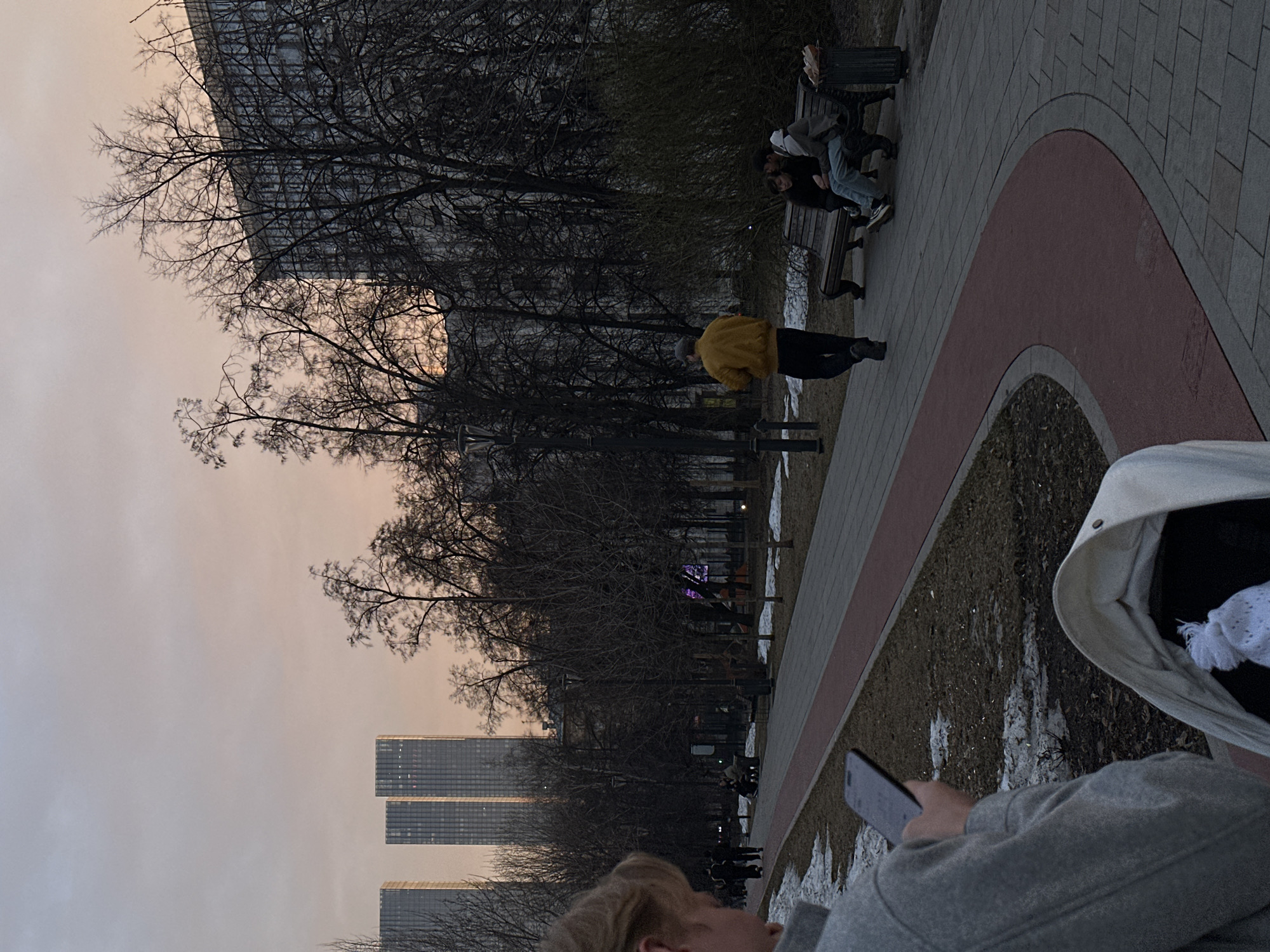 The image depicts an urban park scene in Moscow, Russia, at dusk or late afternoon during late winter or early spring, with patches of snow on the ground and bare-branched trees. In the immediate foreground, a person with light hair, seen from the back-right, wears a grey coat and operates a dark smartphone held in both hands. A light-colored baby carriage or large bag containing a white knitted item rests beside them.
A paved pedestrian path of grey pavers, bordered by a reddish-brown track, extends into the midground. Along this path, a man in a yellow jacket and dark pants walks away from the viewer. Further down the path, two individuals are seated on a dark wooden park bench: one person in dark attire, the other in a grey hooded sweatshirt and light blue jeans. A dark lamppost stands on the left, and a dark trash receptacle is positioned next to the bench.
In the background, numerous bare trees are silhouetted against a pale orange and grey sky. Beyond the trees, several tall, modern glass skyscrapers, characteristic of the Moscow International Business Center (Moscow-City), dominate the skyline. Three distinct rectangular towers are prominent on the left, with another large, multi-story building partially obscured by trees on the right. Distant figures walk further along the park path.