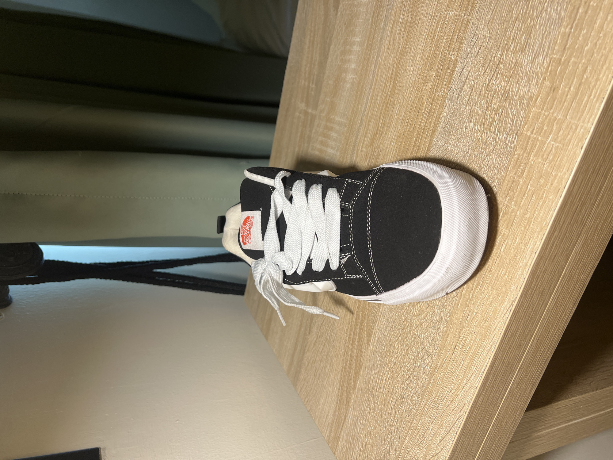 A black and white low-top sneaker is prominently displayed in a vertical orientation against a light brown, wood-grained surface. The shoe features a black suede or canvas upper, crisp white laces tied loosely, and white stitching detailing the panels. A distinctive white rubber toe cap and a ribbed white foxing tape encircles the sole, adding contrast. On the shoe's tongue, a red and white woven label is visible, adorned with stylized white text and a graphic, consistent with a skate shoe brand.
The shoe rests on what appears to be a vertical panel of light-colored wood, characterized by a visible grain pattern running along its length. This wooden surface occupies the right and central portions of the image. To the left of the shoe, the background is dominated by a dark green or olive-toned fabric, possibly a curtain or upholstery, which has subtle horizontal lines and appears somewhat out of focus. Below the main wooden surface, towards the bottom left of the frame, there is a smoother, lighter-toned surface, and a blurred black strap or cable is partially visible.
No discernible activity is taking place; the shoe appears stationary, likely posed for the photograph. The lighting suggests an indoor setting, possibly with natural light from an unseen window or ambient artificial light. There are no specific cues within the image to indicate a precise time of day or weather conditions. The setting appears to be a room within a building. The image contains no visible human subjects or explicit emotional content. The location context provided is Gwarinpa, Nigeria, although there are no specific visual elements within the image to confirm this geographical detail.