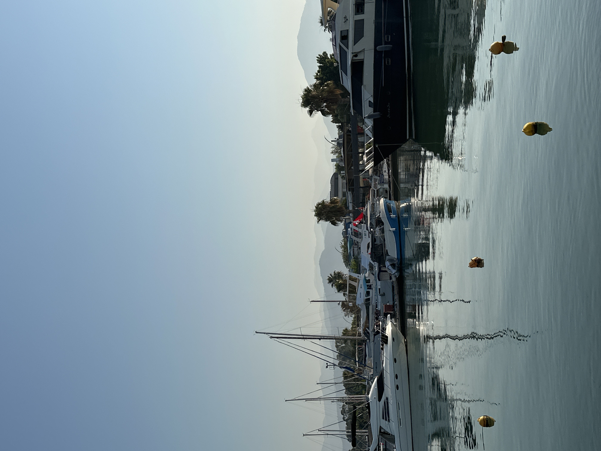 The image captures a serene view of the marina in Fethiye, Turkey, under a clear, pale blue sky. Several boats, including white yachts and sailboats with tall masts, are docked along the waterfront. One prominent dark-hulled yacht, named "Sun of the Sea," is positioned in the foreground on the right. The calm water reflects the boats and the sky, creating a mirror-like effect. In the background, hazy green hills rise, suggesting a coastal mountain range. A few palm trees are visible among the buildings and vegetation along the shore. Yellow buoys float on the water's surface. The scene is tranquil, with no people visible, conveying a sense of peacefulness in the harbor.