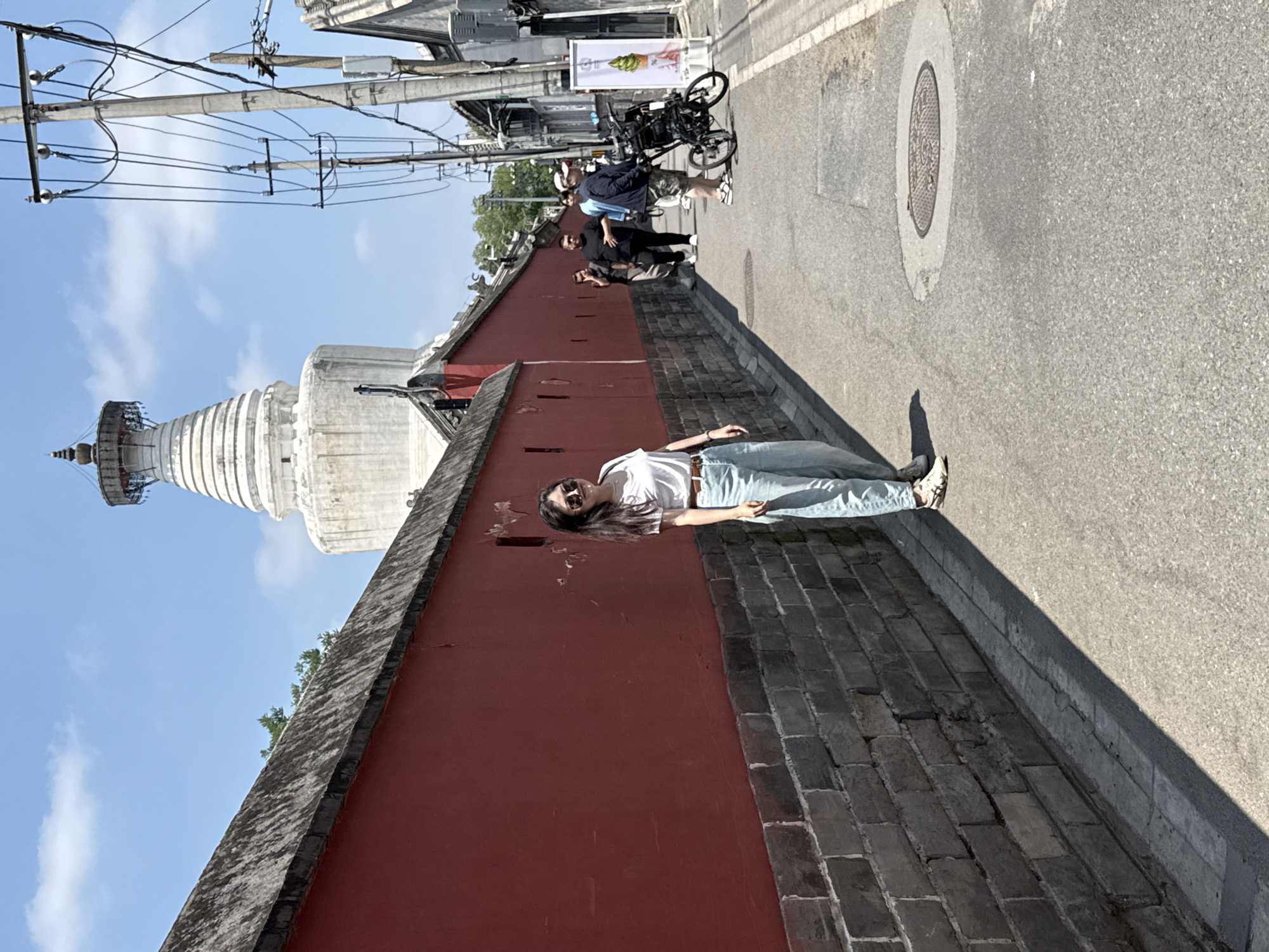 In the Xicheng District of China, a young woman stands on a paved alleyway, leaning against a weathered red brick wall. She wears a white t-shirt, light blue jeans, and sunglasses. The alley is flanked by a high, red-painted wall on the left and buildings on the right, interspersed with utility poles and wires. In the distance, a prominent white stupa with tiered levels rises against a bright blue sky with scattered clouds. Several other people are visible further down the alley, some walking and one with a stroller. A sign displaying an image of green soft-serve ice cream stands near the right side of the alley. The ground is asphalt with a circular manhole cover. The overall scene suggests a sunny day in an urban environment with historical architecture.