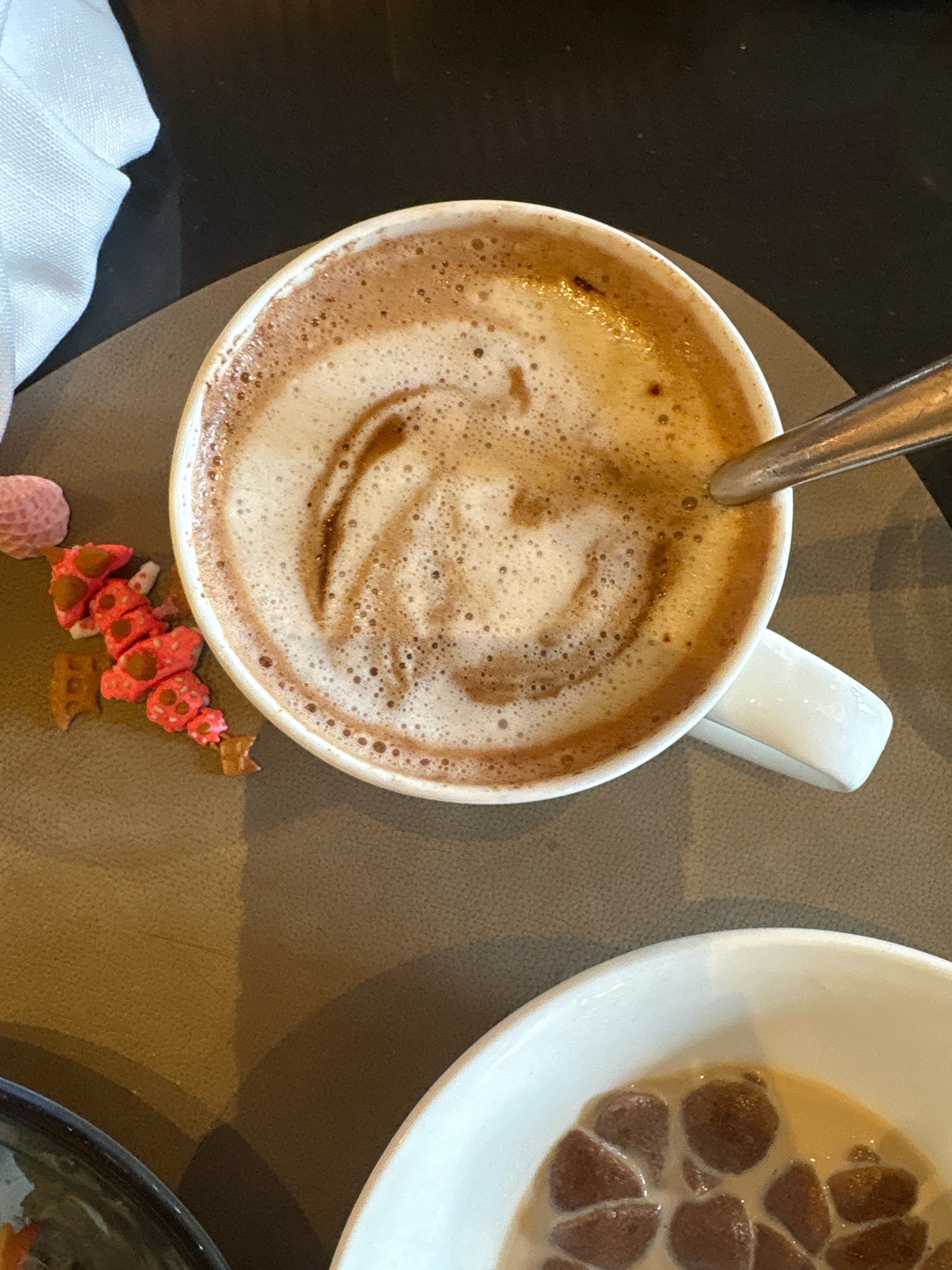 The image shows a close-up, top-down view of a white mug filled with frothy hot chocolate, placed on a dark brown placemat. A silver spoon rests in the mug, partially submerged in the beverage. To the left of the mug, there are small, pink, flower-shaped candies and some broken, square, brown biscuits. In the foreground, a white bowl containing milk and brown, irregularly shaped cereal pieces is partially visible. A white napkin is folded in the upper left corner of the frame, and the background is a dark, textured surface. The scene appears to be indoors, possibly in a cafe or restaurant in Vagator, India.