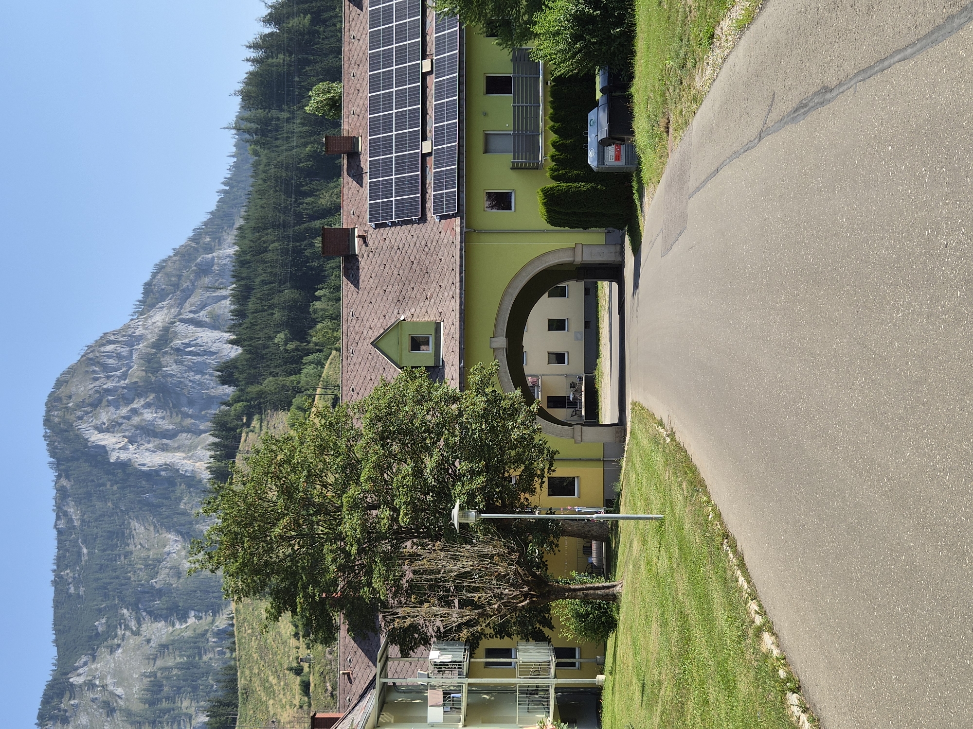 This is a sunny, daytime, eye-level exterior shot of a road leading through an arched entryway of a light green building in Eisenerz, Austria. The building has a brown tile roof with numerous solar panels. To the left of the building is a multi-story structure with balconies. A large green tree stands between the two buildings, with a bare tree to its left. A street lamp is positioned in front of the trees. Behind the building, a forested hillside rises towards a rocky mountain range under a clear blue sky. There are no people visible in the image.