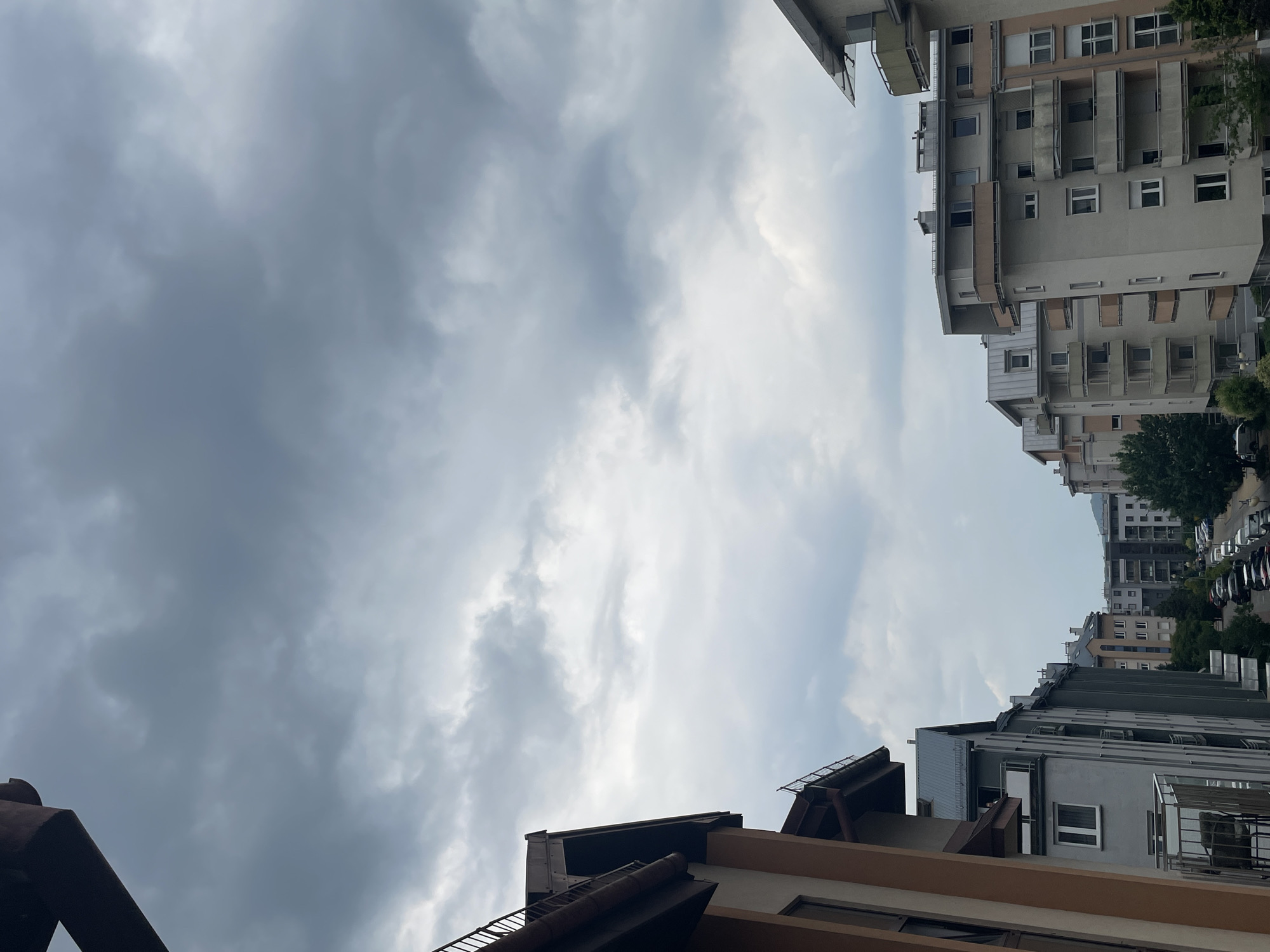 This is a medium shot, eye-level, outdoor, vertical, daytime photograph of a city street scene. The sky is overcast with dark, heavy clouds. In the foreground on the left is the side of a light brown building with a dark brown, sloped roof and a dark brown railing. To the right of this building is a row of multi-story apartment buildings. The buildings are primarily grey and beige with visible balconies and windows. There are trees visible behind the buildings. Further down the street on the right are more apartment buildings and parked cars. The overall atmosphere is somewhat gloomy due to the cloudy sky.