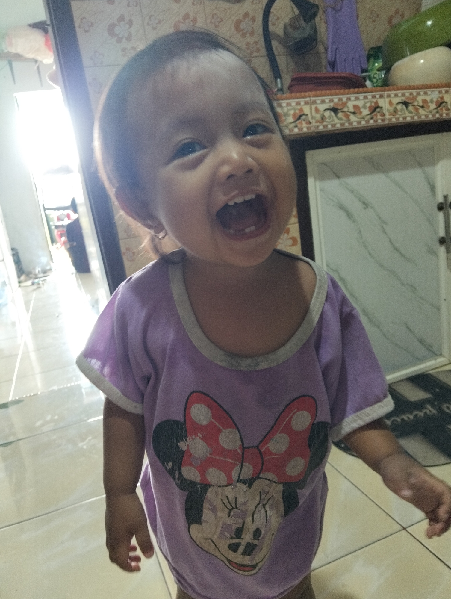 A young child with dark hair and brown eyes is captured in a medium shot, smiling widely with an open mouth, revealing teeth. The child is wearing a purple t-shirt featuring a graphic of Minnie Mouse. The background includes a tiled wall with a floral pattern, a shelf with various items, and a white cabinet. The lighting suggests an indoor setting, possibly a home, with a bright light source to the left of the frame. The scene appears to be in Muncar, Indonesia.