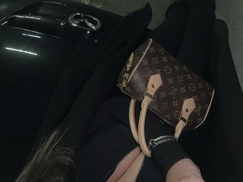 The image depicts a partial view of an individual in a dimly lit environment, identified as Sofia, Bulgaria. The subject, positioned centrally, is wearing a dark, possibly black, garment featuring a prominent vertical zipper detail on the torso. Their light brown hair is visible on the upper left. A silver-colored bracelet adorns their right wrist. A brown Louis Vuitton monogram canvas "Speedy" style handbag, with natural cowhide leather trim and gold-tone hardware, rests on the subject's lap or between their legs. To the right of the subject, the front-right section of a dark-colored Mazda vehicle is visible, displaying the chrome Mazda logo on its grille. Reflections of vertical light sources are evident on the vehicle's glossy surface, consistent with an indoor setting like a parking facility. The immediate ground surface appears light-colored, possibly concrete.