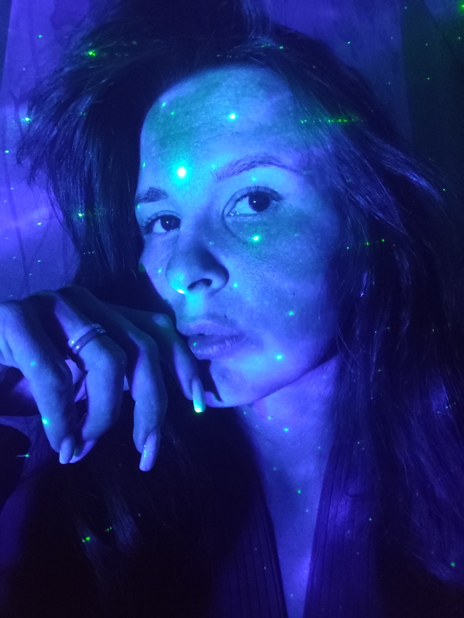 A selfie of a woman with blue and green lights on her face, neck, hair, and hand. The woman is facing the camera, and her hand is resting on her chin. She is wearing a ring on her finger. The background is dark, but it appears to be a room with a curtain in the background. The location is Фоки, Russia.