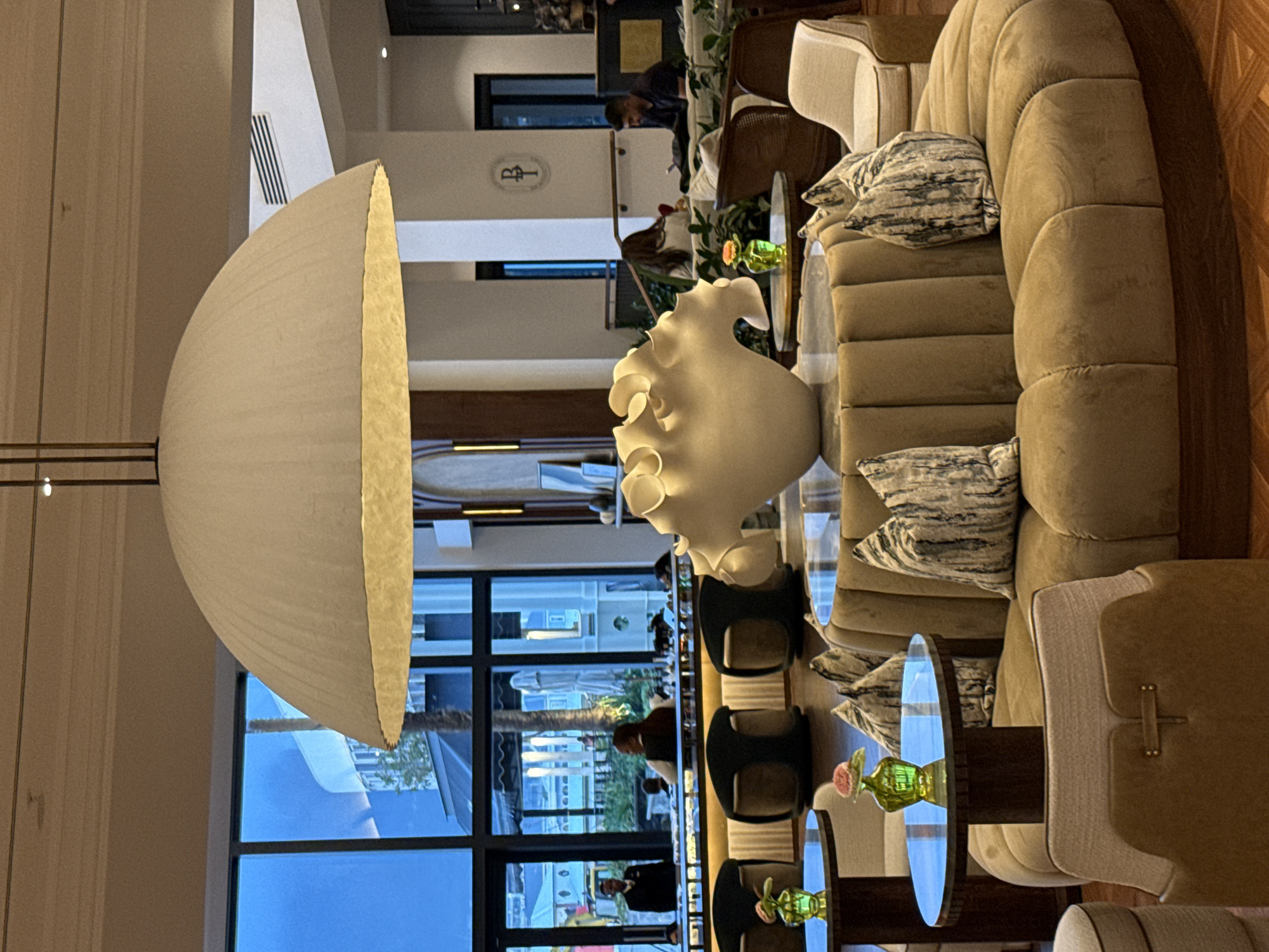 The image depicts a luxurious interior space, likely a high-end hotel lobby or lounge. Features include a large, off-white, dome-shaped lamp fixture dominating the left foreground, partially obscuring the background. To the right is a large, curving sofa with patterned pillows. Glass-topped tables with floral arrangements are arranged near the seating. Through large windows, a partial view of a city environment with a possible outdoor seating area is visible. The architecture suggests a contemporary design aesthetic. People are present in the scene, engaged in activities consistent with a lobby environment. The location is identified as Cape Town, South Africa based on the architecture.