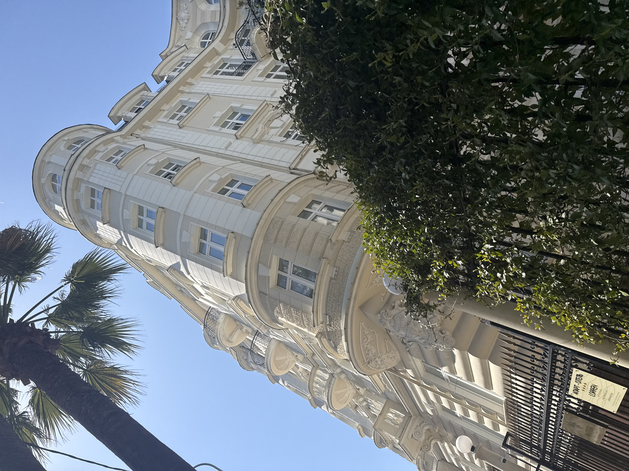 The image shows a view of a building in Unknown, Unknown. The building has a light-colored facade and numerous windows. A portion of the building is rounded, like a tower. There is a tree partially obscuring the building on the right side of the frame and a palm tree on the left side. A metal fence is visible at the bottom right, with a sign that reads "LOREN" in Russian script. The sky is a clear, light blue.