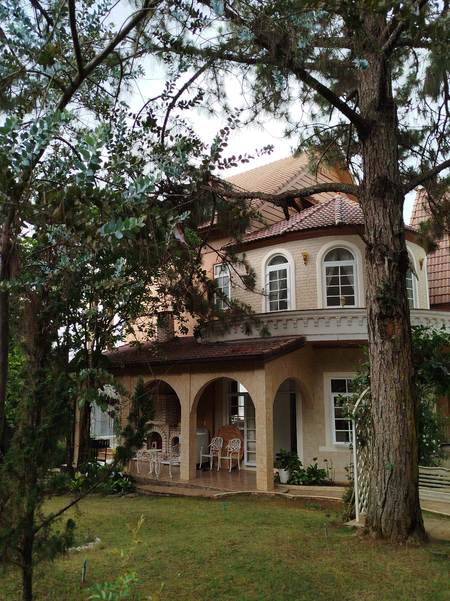 The image depicts a two-story house in Da Lat, Vietnam. The house has a cream-colored facade, arched windows on the second floor, and a red-tiled roof. A covered porch with arched supports runs along the front of the house, furnished with white wrought-iron furniture. A brick fireplace is visible at one end of the porch. A well-maintained lawn stretches out in front of the house, and the property is surrounded by lush trees, partly obscuring the view of the house.
