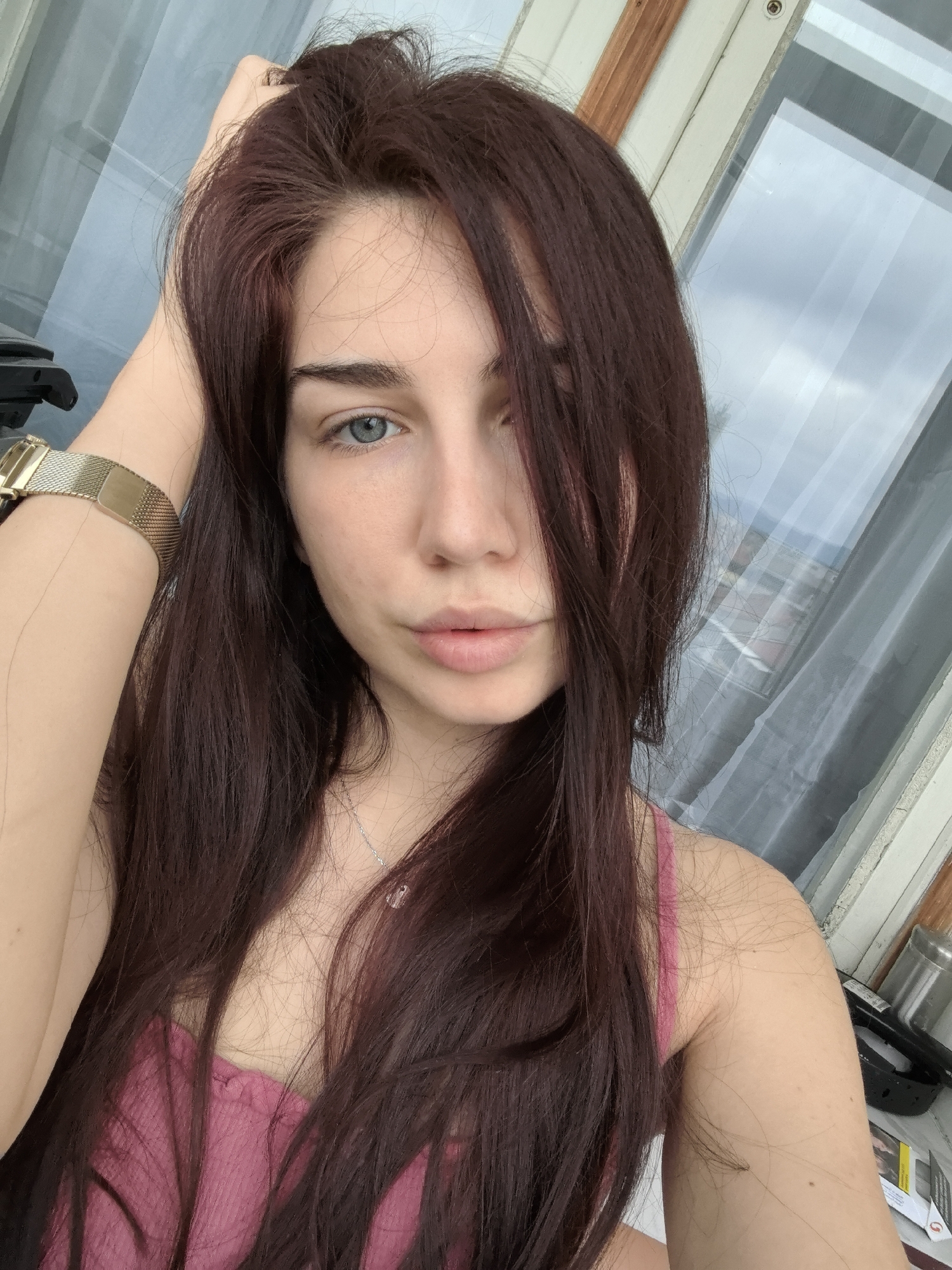 A young woman with long, dark auburn hair stands indoors, likely in an apartment, based on the window with a glimpse of an urban landscape. She wears a pink tank top and a gold watch on her left wrist. Her hand is raised towards her hair. She looks directly at the camera with a slightly pouty expression. The blurred background suggests a cityscape, possibly Stara Zagora, Bulgaria.